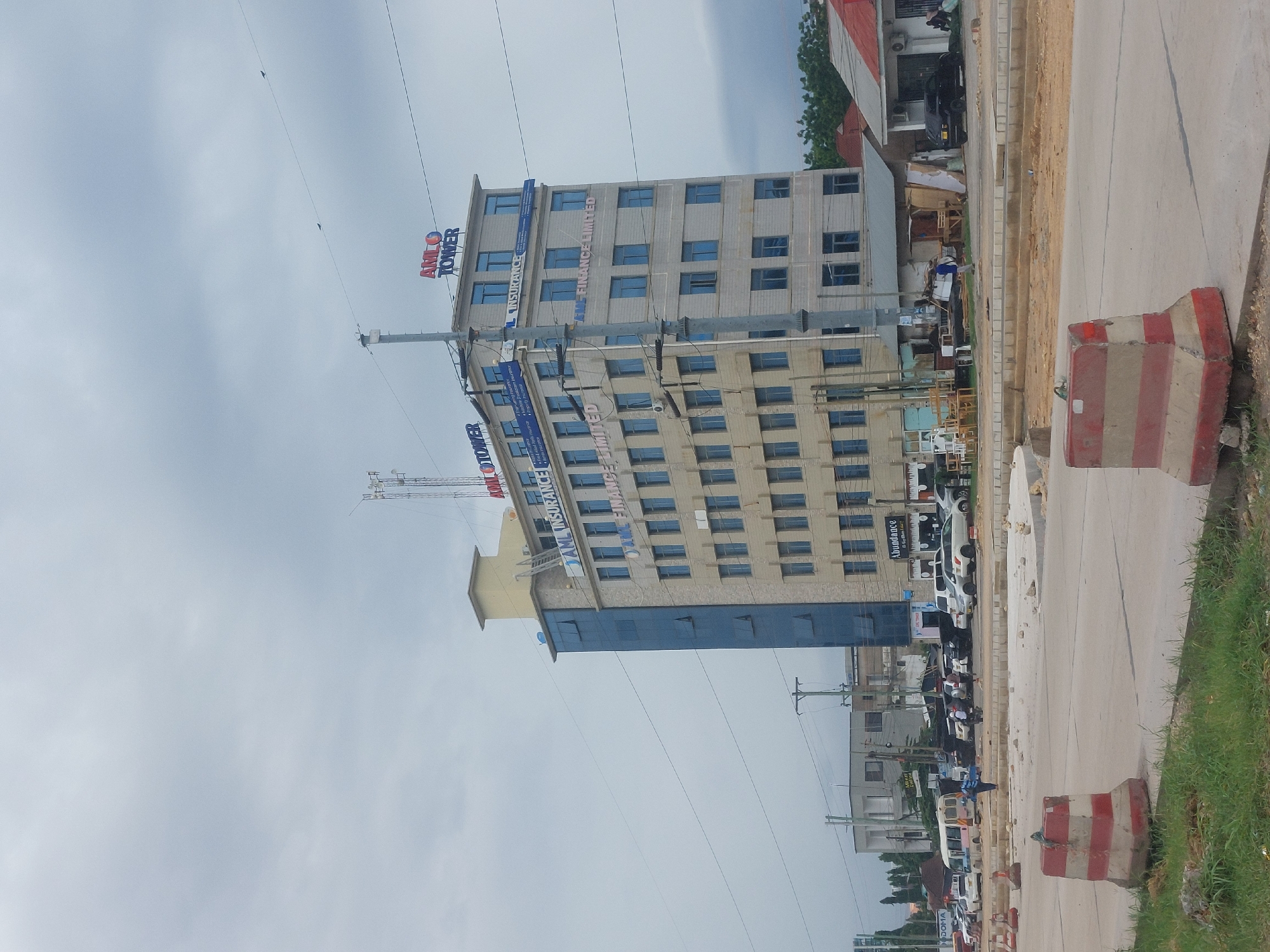 The image depicts a multi-story building, identified as "AMIL Tower," in Dar es-Salaam, Tanzania. The building has a combination of grey and blue facade elements. Street-level traffic and infrastructure are visible in front of the building. A section of roadway with painted barricades is also present. The sky is overcast.