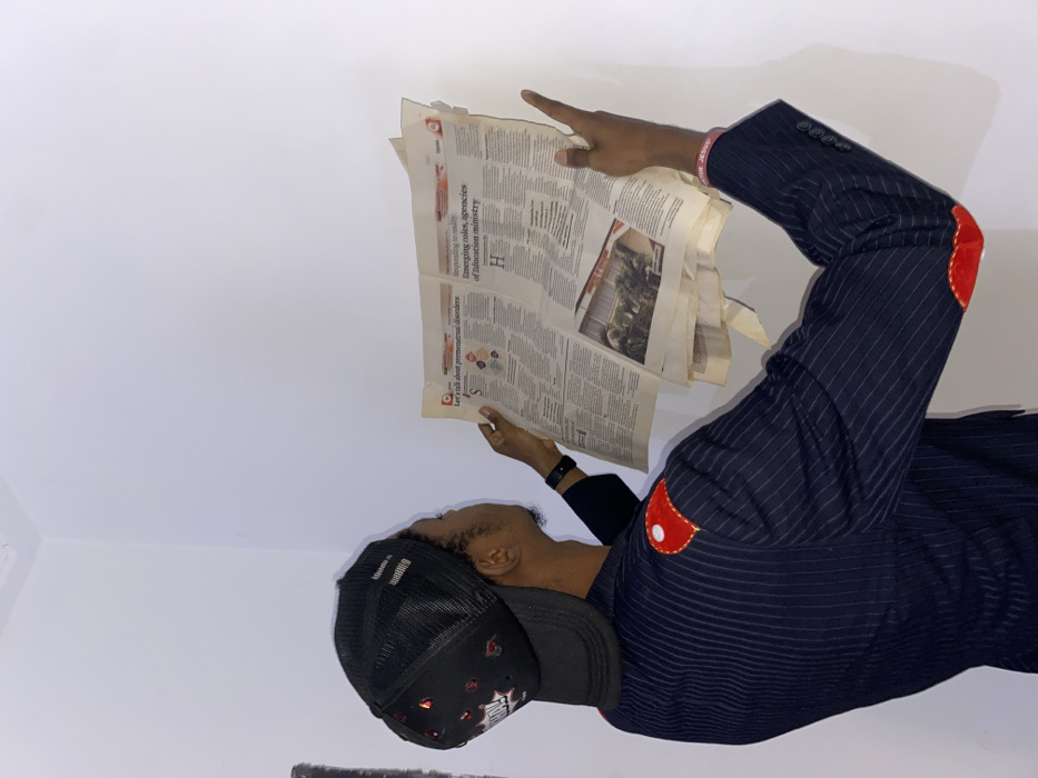 An individual is depicted reading a newspaper, seen in a rotated orientation with their head facing left. The person, who appears to be male, is wearing a black mesh baseball cap and a dark pinstriped jacket adorned with distinctive red, circular elbow patches. A black wristband is visible on their left wrist, and a pink wristband with the text "FOR JESUS" is on their right arm. They hold an open newspaper with both hands, and their right index finger is pointing at a section of the page. The background is a plain, light-colored wall or surface, indicating an indoor setting.
Visible text on the newspaper includes headlines such as "Let's talk about premenstrual disorders", "Responding to reality", and "Emerging roles, agencies of Education ministry". There is also a large "H" and "0000" visible. The person's expression is not fully clear due to the angle, but they appear focused on reading. The scene takes place in Joma, Ghana.