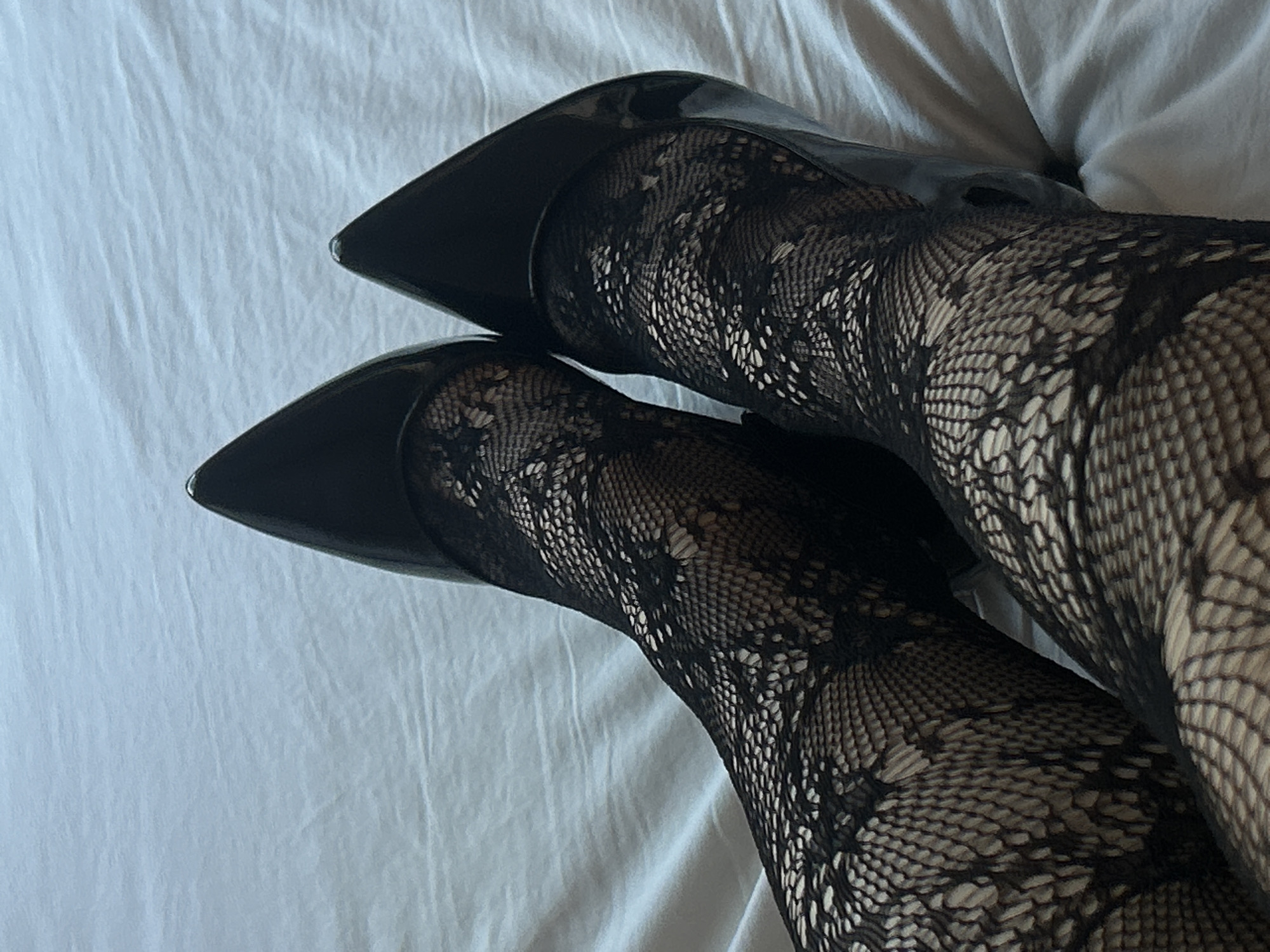 The image shows a pair of legs clad in patterned black tights and black patent leather high heels resting on a white surface. The feet are crossed at the ankles. The background is a crumpled white cloth, possibly a bed sheet. The location is not discernible from the image and cannot be identified as Novi Sad, Serbia or any other location.