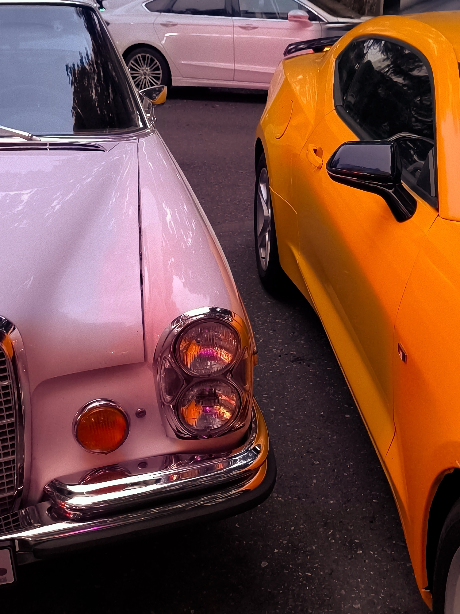 A close-up, low-angle shot captures the front of a light pink vintage car and the side of an orange modern sports car parked side-by-side on a dark asphalt surface. The vintage car's chrome grille and dual round headlights are prominent, with a red indicator light positioned below the headlights. The orange sports car's sleek body, black side mirror, and wheel are visible. In the background, a white modern sedan is parked. The scene appears to be set in Batumi, Georgia, likely on a street or parking area, as suggested by the overall urban environment.