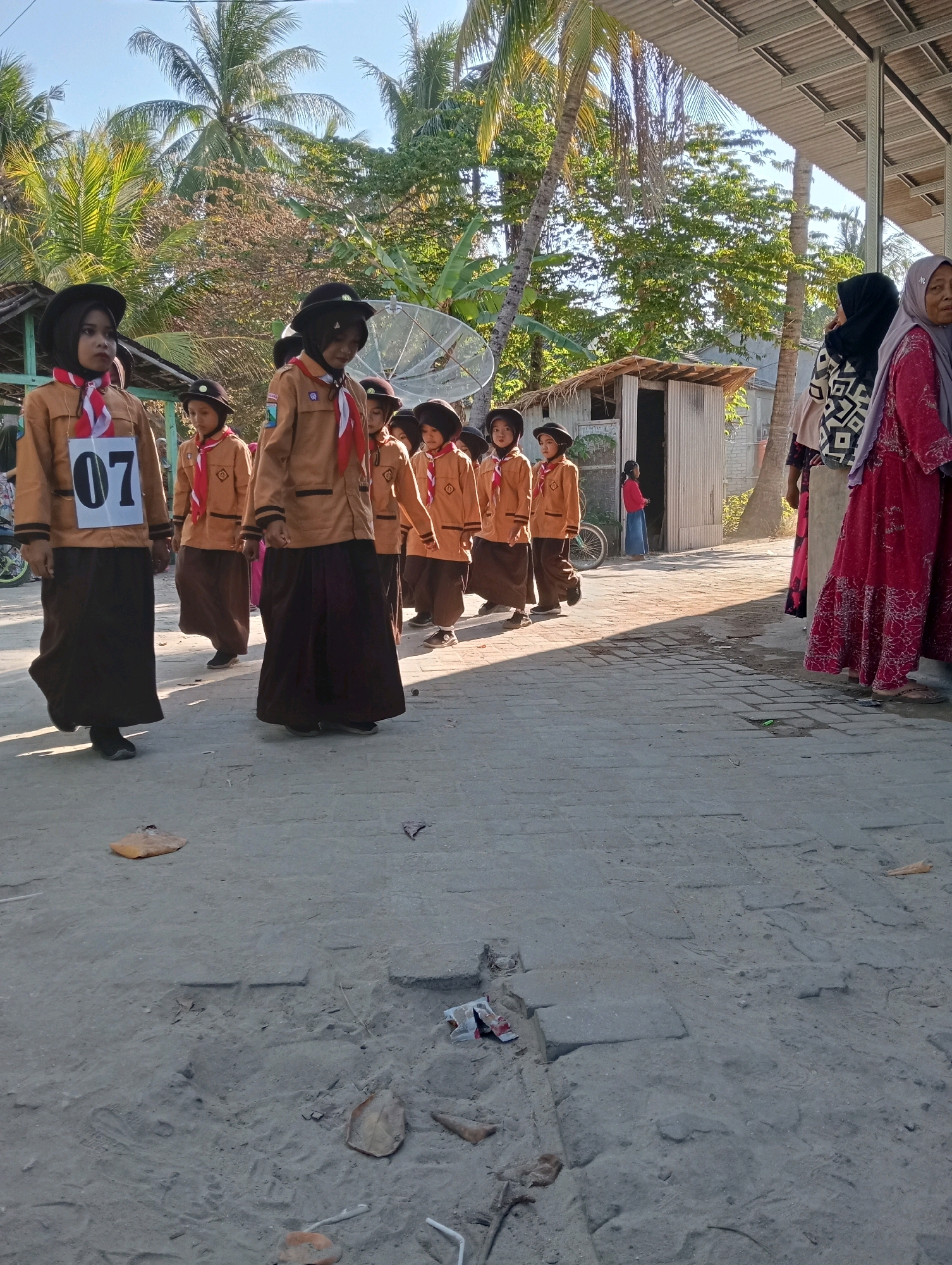 A group of young girls in uniform are walking down a dusty path in what appears to be a village in Pagerungan Kecil, Indonesia. The girls are dressed in brown uniforms with red and white scarves, and some are wearing black hats. One girl in the foreground has a large white number "07" pinned to her uniform. The path is lined with palm trees and some simple buildings. In the background, two women are standing near a building, and a child in a red shirt is standing in a doorway. The sun is shining, casting long shadows on the ground.