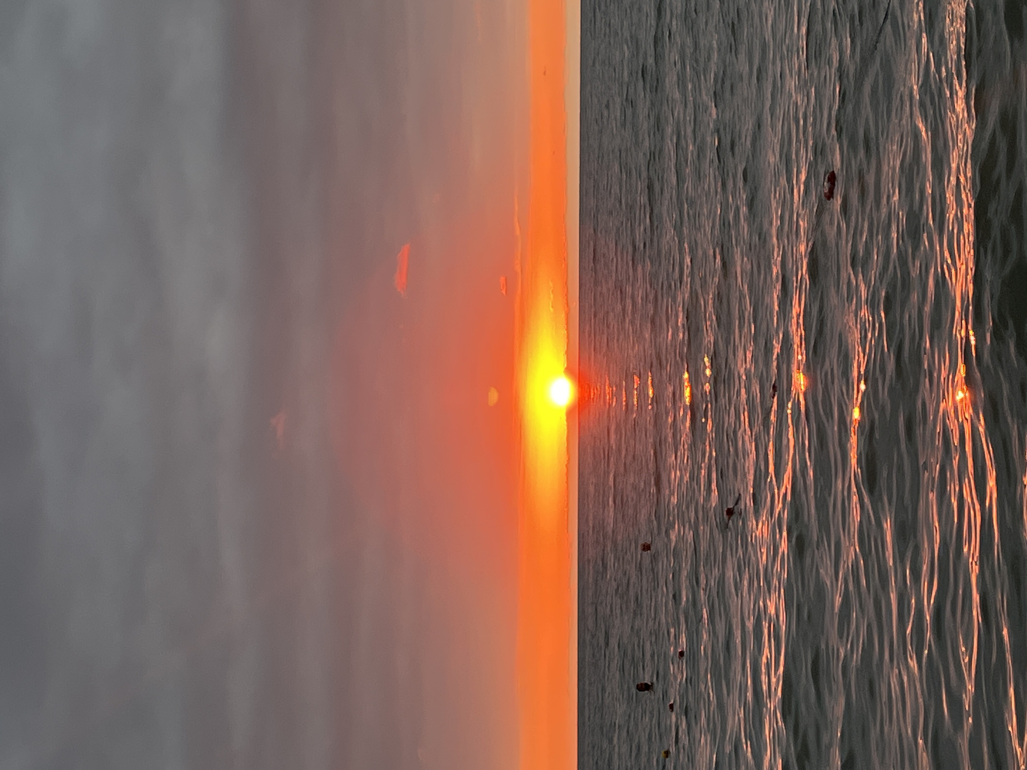 The image depicts a serene sunset over the Black Sea in Ureki, Georgia. The sky is painted with vibrant hues of orange and red, transitioning to a dusky gray at the top, with a few wispy clouds scattered across. The sun, a brilliant orb of orange, is partially obscured by the horizon, casting a shimmering golden path across the rippling water. Several buoys dot the sea, suggesting a designated swimming area. In the foreground, the dark blue water is illuminated by the sunset's glow, with subtle waves reflecting the warm light. Two individuals are visible in the water, their heads just above the surface, appearing to swim or relax in the sea. The overall impression is one of peaceful tranquility as the day draws to a close.