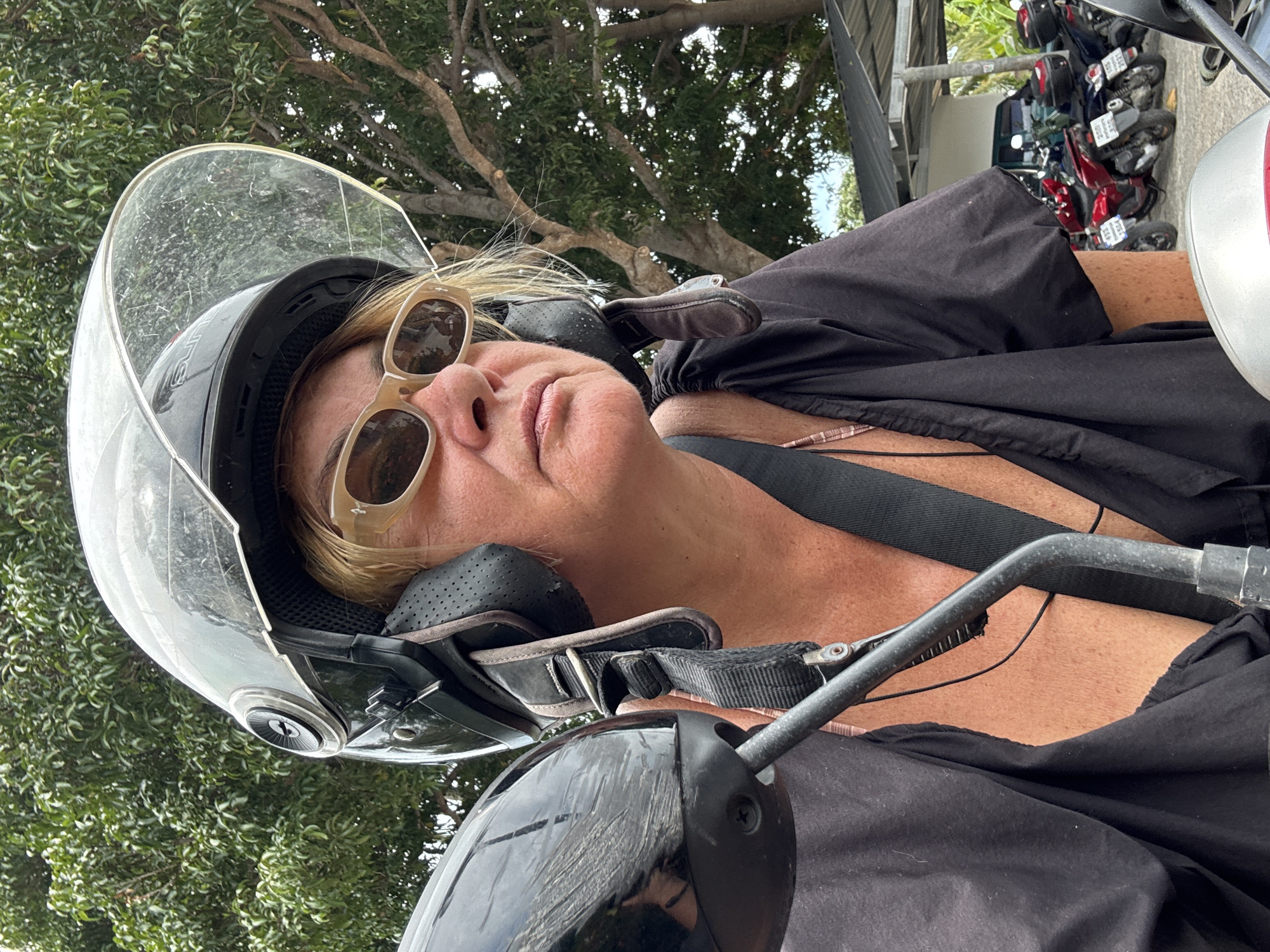 A woman wearing sunglasses and a motorcycle helmet is riding a scooter in Baan Tai, Thailand. The environment includes trees, other parked motorcycles, and a building.