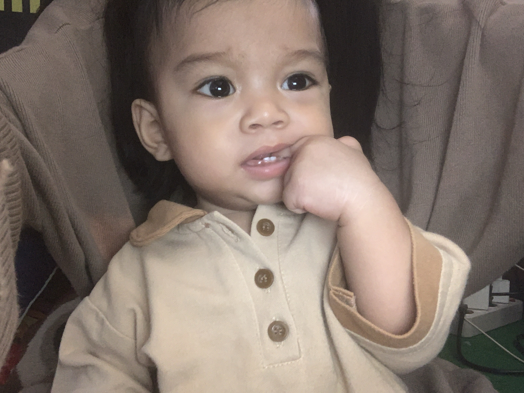 A close-up, head-and-shoulders shot focuses on a young child, appearing to be less than two years old, sitting and possibly being held. The child has dark hair and large, dark eyes. One hand is raised to the child's mouth, with fingers touching the lips and visible teeth. The child is wearing a light brown collared shirt with three visible brown buttons down the front. The background is soft and out of focus, showing textures of fabric, possibly a sweater or blanket, in neutral tones of brown and beige. The overall lighting is dim. The location in Sungai Meriam, Indonesia, is not discernible from the visual information in the image.
