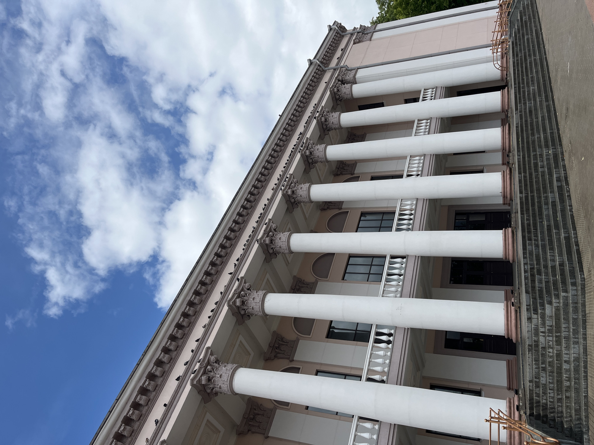 The image shows a wide shot of the building housing the Hrodna Regional Drama Theatre, located in Hrodna, Belarus. The building features a classical facade with a colonnade of white Ionic columns supporting a light pink entablature. A white balustrade runs along the second-story balcony. The sky above is a bright blue with scattered white clouds. Several pigeons are perched on the decorative molding along the top of the building. In the foreground, a wide set of stone stairs leads up to the entrance. The overall impression is one of stately architecture under a clear sky.