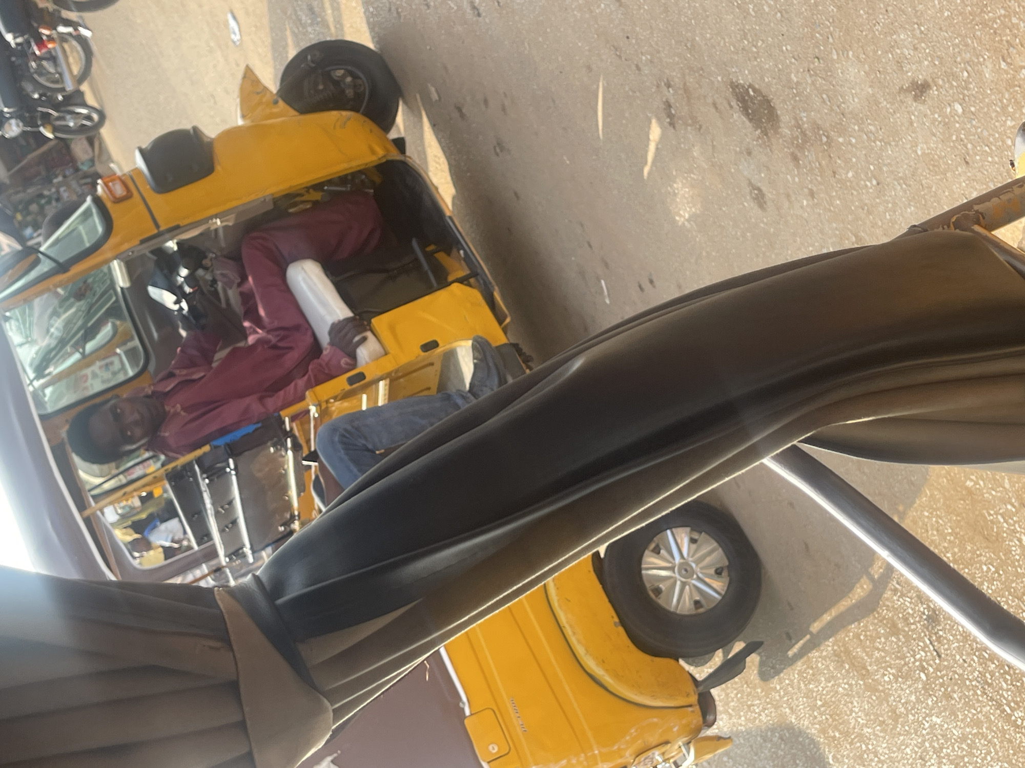 In Galadimawa Village, Nigeria, a young boy, dressed in a maroon kaftan and blue jeans, sits at the helm of a yellow auto-rickshaw, likely engaged in its operation or in transit. The vehicle, a common mode of transport in the region, is partially obscured by the foreground, which appears to be the interior of another vehicle or structure, possibly a canopy or a seat, covered in dark fabric with a beige trim. The scene unfolds on a dusty, sun-drenched street, with the bright sunlight casting distinct shadows. In the background, other auto-rickshaws and motorcycles suggest a busy urban or semi-urban environment. The boy's gaze is directed forward, hinting at his focus on the road ahead.