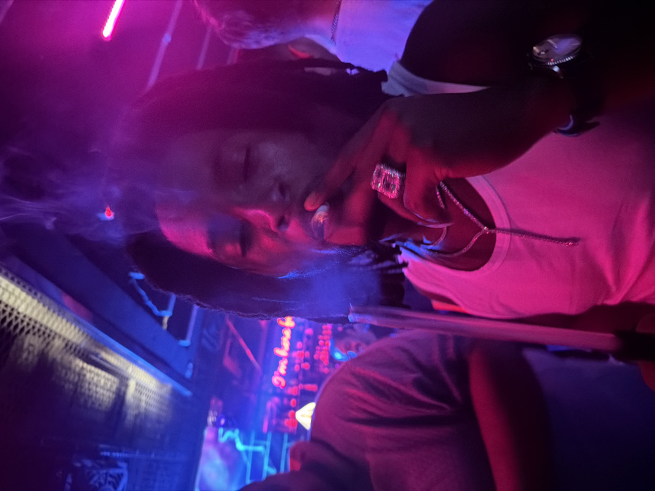 A person with dreadlocks is smoking, creating a cloud of smoke around their face. They are wearing a white tank top and a large diamond ring on their finger. A diamond necklace is also visible. The scene is lit with bright pink and blue neon lights, creating a club-like atmosphere. A sign that reads "I'm here for" is visible in the background.