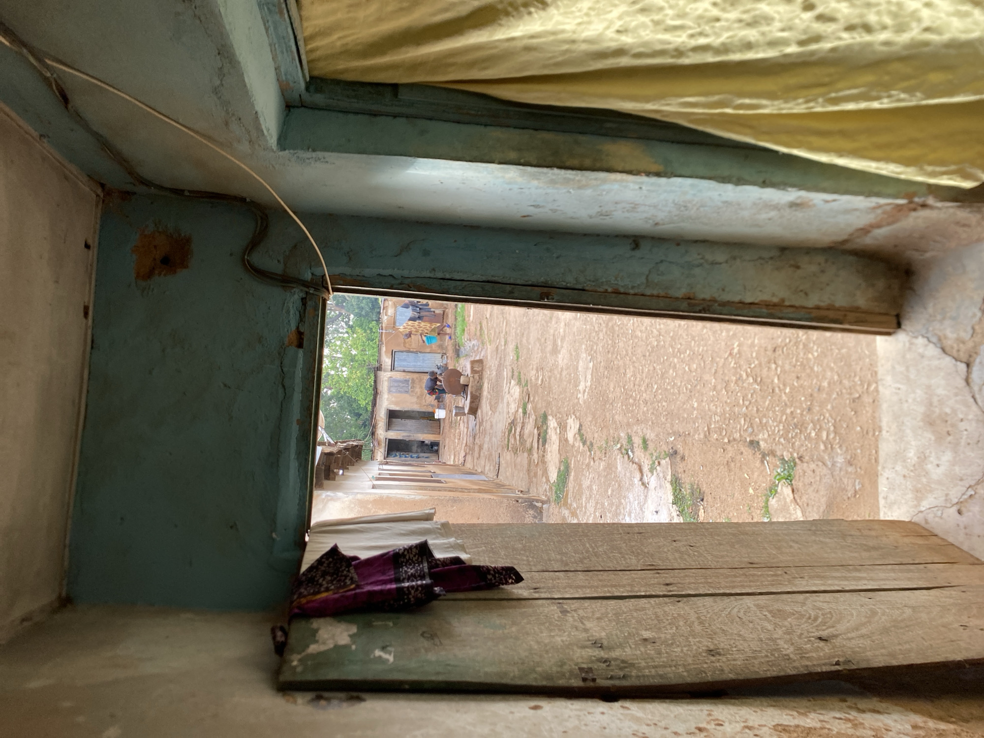 The image shows a view looking out from a room or space onto a street in Bukuru, Nigeria. The foreground is dominated by a rough, wooden surface, possibly a windowsill or a partially constructed platform. A folded piece of purple and black patterned fabric rests on the edge of this wooden structure. The walls of the room are painted a faded teal color, showing signs of wear and age. An electrical wire is visible, running along the wall near the window frame.
The view through the opening reveals a street or courtyard area with a packed-earth surface. There are buildings visible in the distance, with people performing various activities, including what appears to be cooking with open fire. The overall impression is of a simple, rural or semi-urban environment.