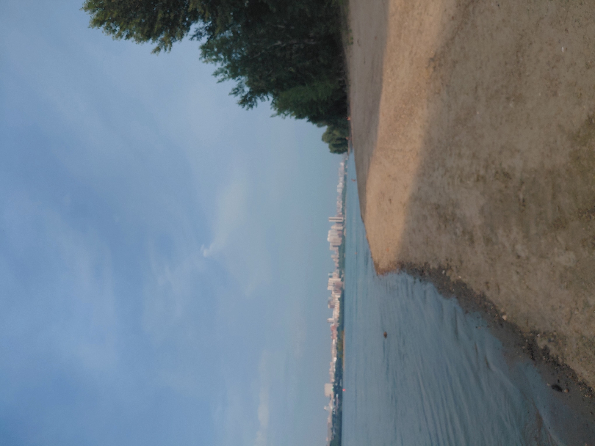 The image shows a view of the Ob River on a clear day, with a beach in the foreground and the cityscape of Novosibirsk, Russia, in the background. The beach is sandy and appears to be unoccupied, with the gentle waves of the river lapping at the shore. Lush green trees line the riverbank to the right of the image, providing a stark contrast to the urban skyline on the opposite bank. The cityscape features a mix of modern buildings and older structures, suggesting a vibrant and developed urban center. The sky is a soft blue, with a few wispy clouds adding texture to the scene. The overall impression is one of a tranquil and picturesque day at the river, offering a glimpse of the natural beauty and urban life of Novosibirsk.