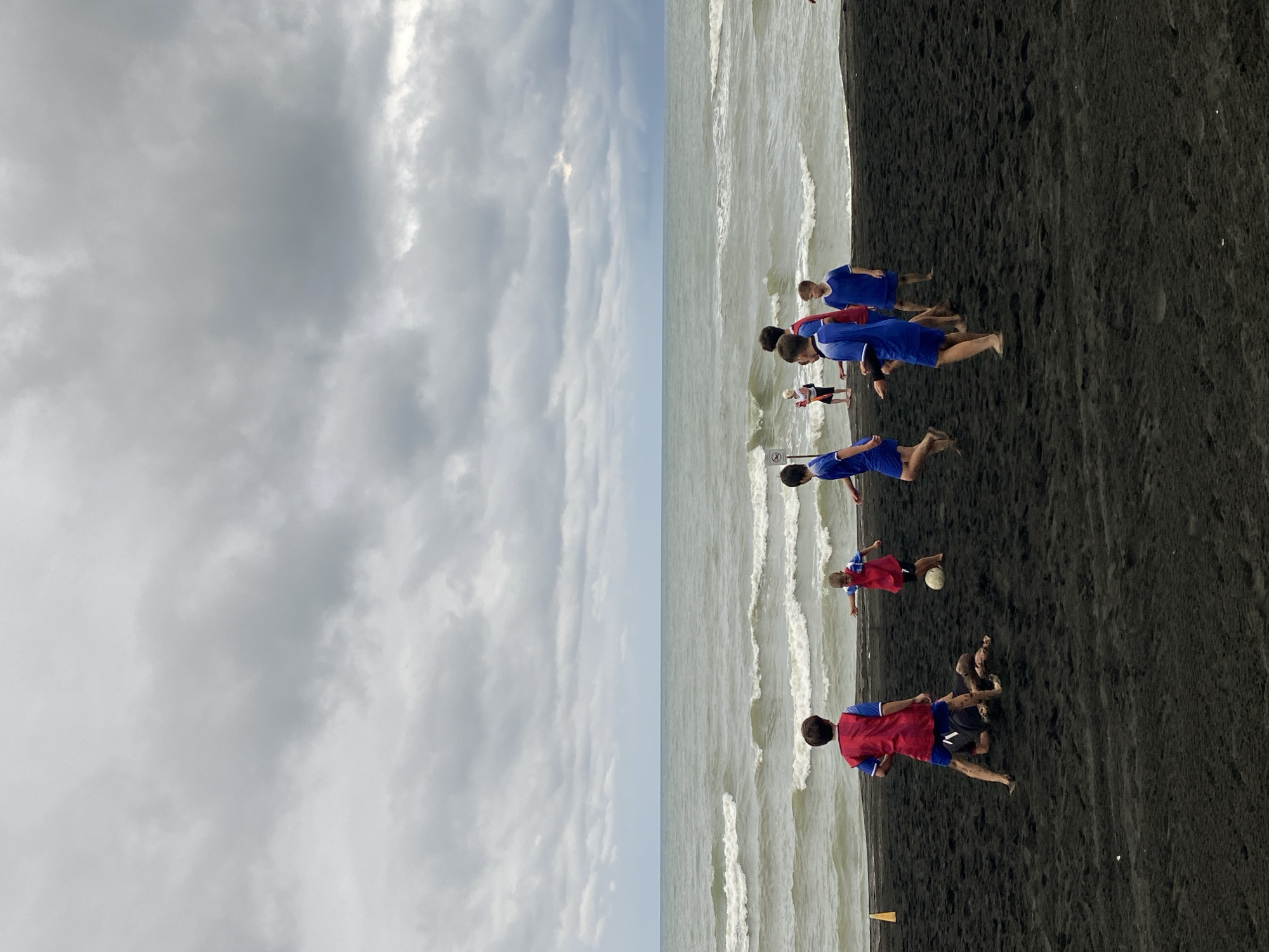 The image depicts a group of children playing soccer on a black sand beach in Ureki, Georgia. The sky is heavily overcast with thick, gray clouds, suggesting potential for rain. The sea in the background is choppy with visible waves. On the beach, several children are actively engaged in the game. One child in a red vest is positioned towards the left of the frame, facing away from the camera. To his right, another child in a red vest is kicking a soccer ball. Several other children, some in blue and red attire, are scattered across the sandy expanse, participating in or observing the game. One child is lying on the sand near the center of the image. The black sand beach is characteristic of Ureki, known for its therapeutic magnetic sand.
