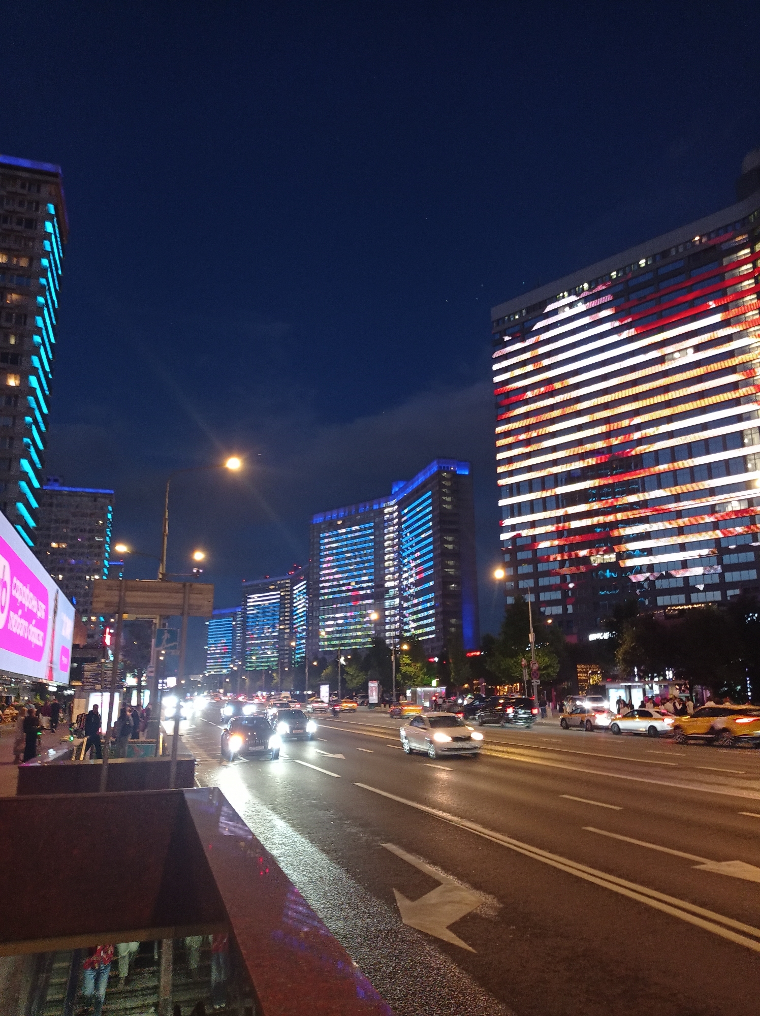 This is a nighttime photograph taken in Moscow, Russia, likely on Kutuzovsky Prospekt, a major avenue known for its illuminated architecture. The scene depicts a wide, multi-lane road with several cars driving on it, their headlights and taillights creating streaks of light due to the long exposure. The road surface is wet, reflecting the lights.
On the right side of the road stands a prominent, tall building with a curved facade. Its exterior is illuminated with a dynamic light display featuring horizontal stripes of white and red, creating a striking visual effect. Further down the road, on the same side, are more tall buildings, also lit up with blue lights outlining their structures.
On the left side of the road, another tall building is visible, its facade lined with bright blue lights. In the foreground, a pedestrian walkway or entrance to an underground passage is visible, with a dark, red-brown marble-like material forming its walls. Several people are seen on the sidewalk and near the entrance, some of them illuminated by the bright pink billboard on the far left. The sky above is a deep, dark blue, characteristic of a city at night. The overall atmosphere is urban and vibrant, showcasing Moscow's modern architecture and illuminated cityscape.