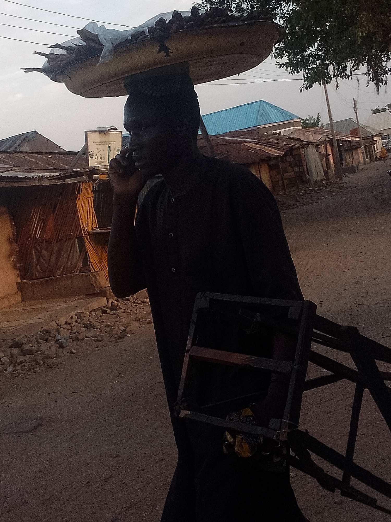 In Pantame, Nigeria, a person walks along a dirt road. They carry a tray of goods on their head and a wooden structure. They are using a mobile phone. Background consists of corrugated metal buildings and other signs of settlement.