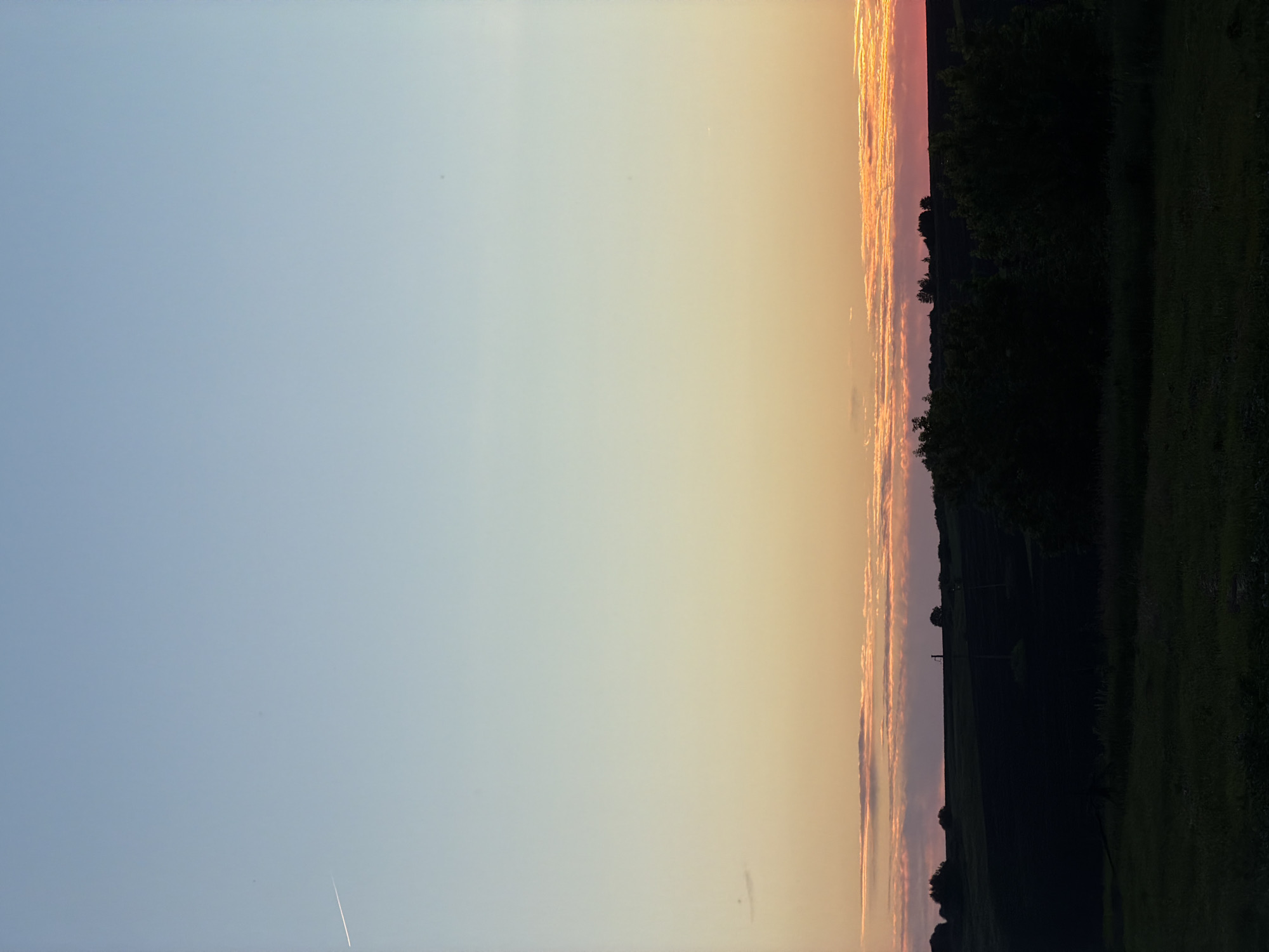 The image depicts a rural landscape at sunset. The sky is a gradient of soft blues and yellows, transitioning to vibrant oranges and pinks near the horizon, indicative of the time of day. Long, wispy clouds are illuminated by the setting sun, creating a dramatic effect.
The foreground is dominated by a dark silhouette of trees and rolling hills, suggesting a wooded or forested area. The contrast between the bright sky and the dark land creates a sense of depth and atmosphere. A small, faint contrail is visible in the upper left quadrant of the sky.
The scene evokes a feeling of peace and tranquility, common emotions associated with sunsets. The location cues, such as the type of vegetation and the rolling hills, are consistent with a rural setting in Petryaksa, Russia. There is no visible text in the image.