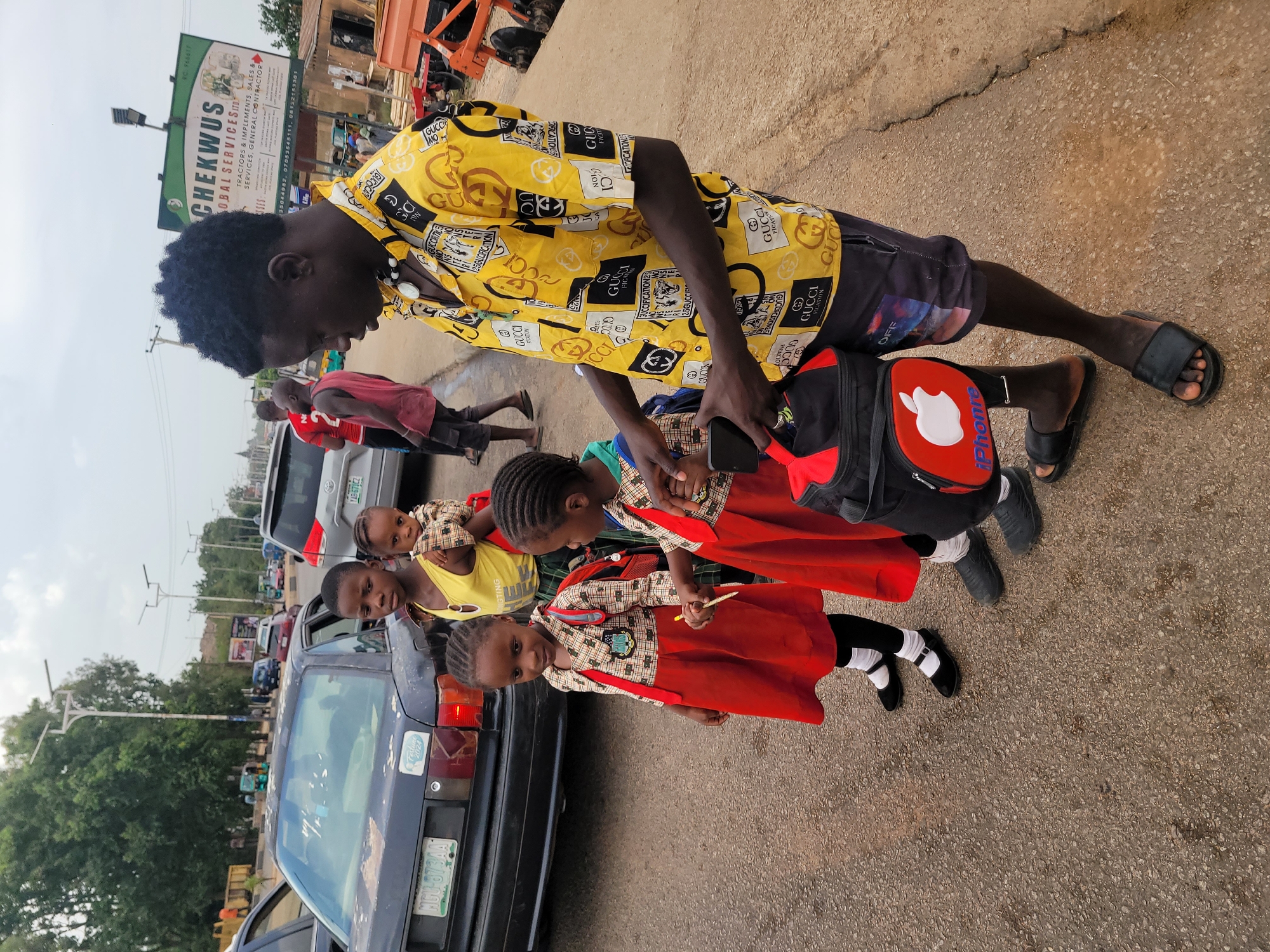 The image captures a street scene in what appears to be a commercial area of Jos, Nigeria. In the foreground, a young man wearing a yellow Gucci print shirt, black shorts, and sandals is bending over, looking into a red and black backpack with a white apple logo on it. He is holding a black smartphone. Four young girls are standing next to him. Three of them are wearing matching red school uniforms, consisting of red dresses with plaid tops. The uniform is completed with white socks and black shoes. The fourth child is wearing a yellow tank top. They are standing next to a dark-colored car. In the background, there is traffic, other pedestrians, buildings, and a billboard advertising "CHEKWUS GLOBAL SERVICES LTD" for tractors and implements sales and services. The environment is paved and appears to be a busy roadside with various parked vehicles and other people in the periphery. A license plate is visible on the dark car.