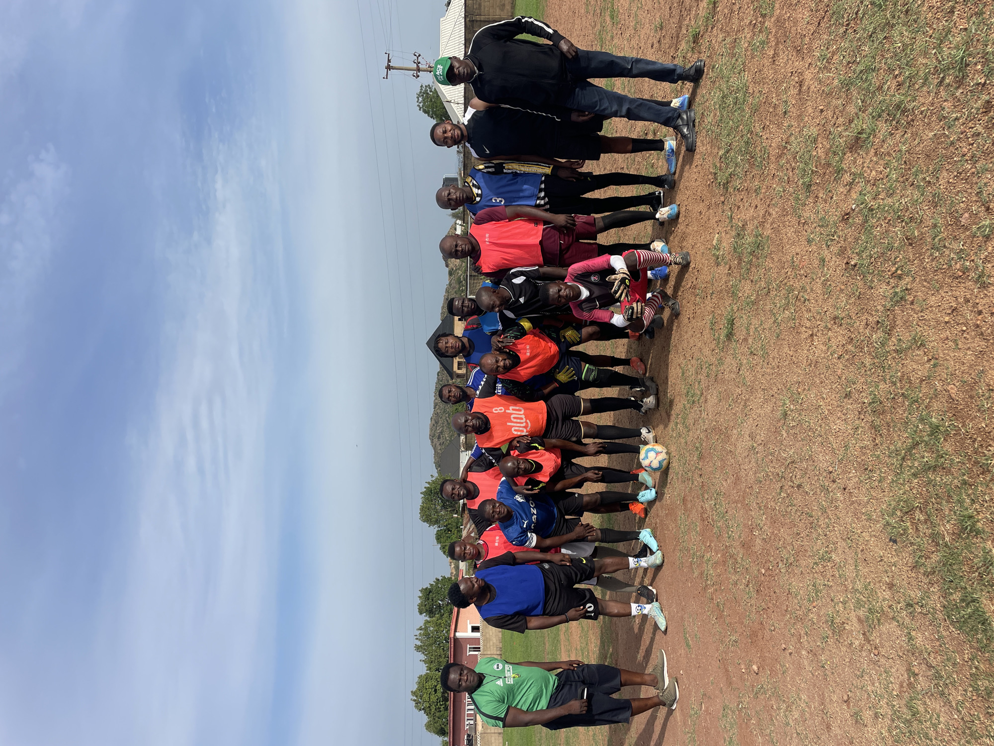 A group of approximately 18 individuals are lined up in a casual team photo setting, on what appears to be a dirt field, near the city of Jos, Nigeria. The sky is light blue with sparse clouds. Behind the group, there are some buildings and what looks like a power line. The individuals are wearing different sportswear, with some in matching colored jerseys and shorts, suggesting they are likely a sports team. A soccer ball is visible amongst their feet. One individual is standing away from the main group, wearing a green t-shirt and black shorts. Several of the individuals are wearing gloves.