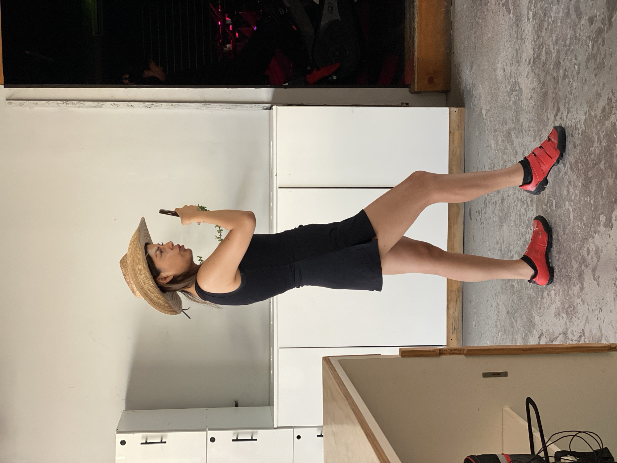 An indoor, vertical full-body shot shows a woman standing, looking down at a smartphone held in her left hand. She wears a straw hat, a black sleeveless mini-dress or romper, black socks, and bright red athletic shoes with black soles and three straps, resembling cycling shoes. Her long, light brown hair is visible under the hat, and a small green plant is held near her left hand.
The setting appears to be a modern fitness studio or gym. The background features a large dark area with exercise equipment, which includes what looks like spinning bikes with red accents and partially visible text, possibly "BA," on one of the machines. Below this equipment are sleek white storage cabinets or lockers with black handles, set against white walls. The floor is a light gray concrete with textured markings. A wooden counter or table is visible in the lower right foreground. The lighting suggests an indoor, artificial light setting. The overall mood is casual and focused, with the woman engaged with her phone. The location context is Puebla, Mexico.
