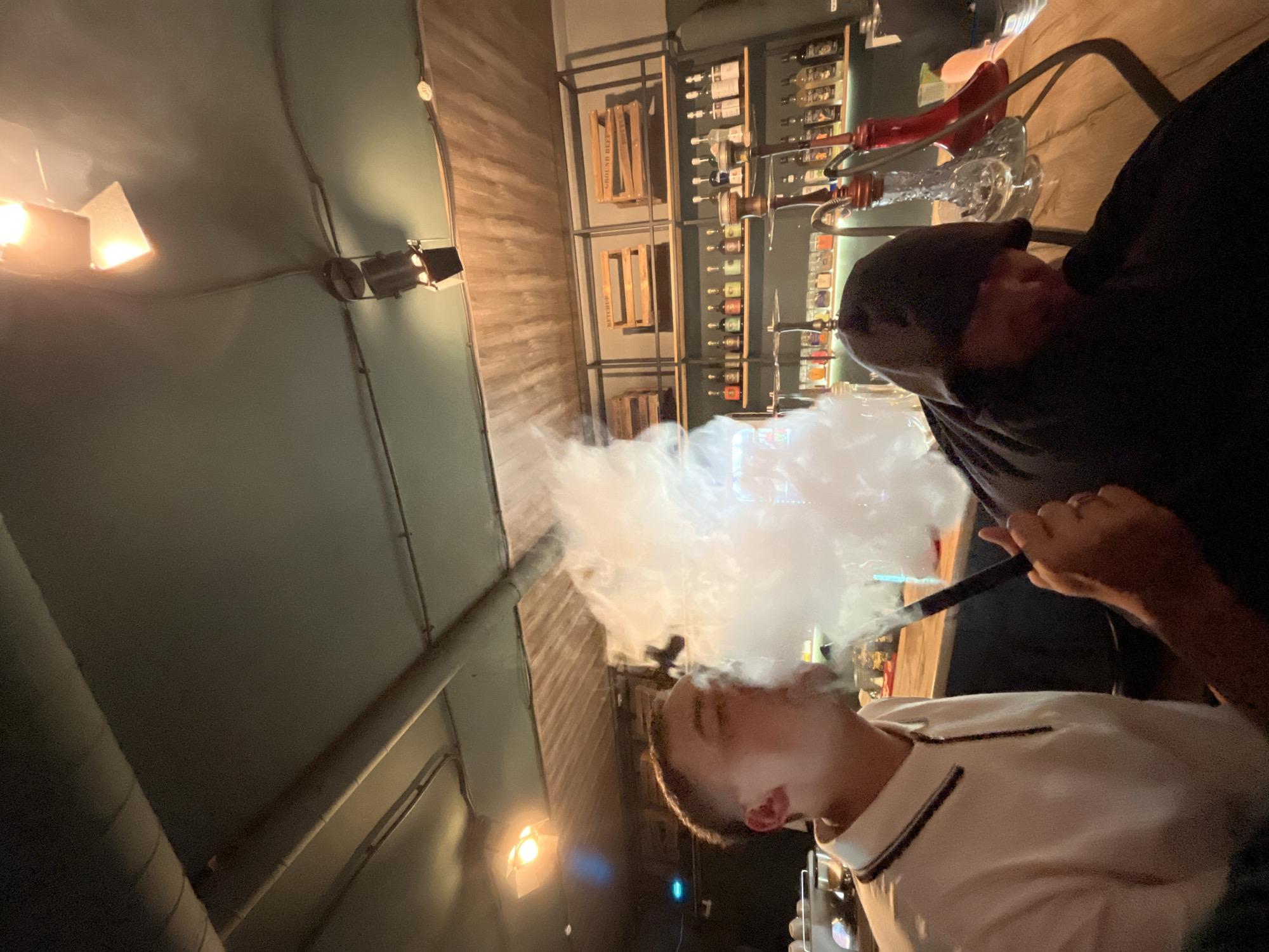 The image depicts an interior scene within a commercial establishment, identified as a hookah lounge or bar located in Chaikovsky, Russia. Two male individuals are positioned at a light-colored wooden counter. The individual on the left, wearing a white short-sleeved polo shirt with dark trim, is actively exhaling a significant cloud of white vapor from a hookah, holding the dark hose mouthpiece with his right hand. His profile and short dark hair are visible, partially obscured by the vapor. The individual on the right is largely obscured by a dark, possibly hooded garment; he appears to be in the process of smoking a hookah, with a dark mouthpiece positioned near his face.
In the background, a metal shelving unit is mounted against a textured wooden wall. The upper shelves display several light-colored wooden crates, two of which are labeled "KETTCHUP" and "GROUND BEEF." The lower shelves are stocked with numerous bottles, consistent with alcoholic spirits or mixers, and are illuminated from below by strip lighting. Multiple hookah bases, including clear glass and red-tinted models, are visible on the wooden counter and the shelves, some with attached hoses. The ceiling is dark, featuring exposed pipes and conduits, along with several track-mounted spotlights that provide directed illumination to specific areas of the room. A blue light source is visible in the distant left portion of the image. The overall environment suggests a dim, lounge-like ambiance with an industrial-chic design.