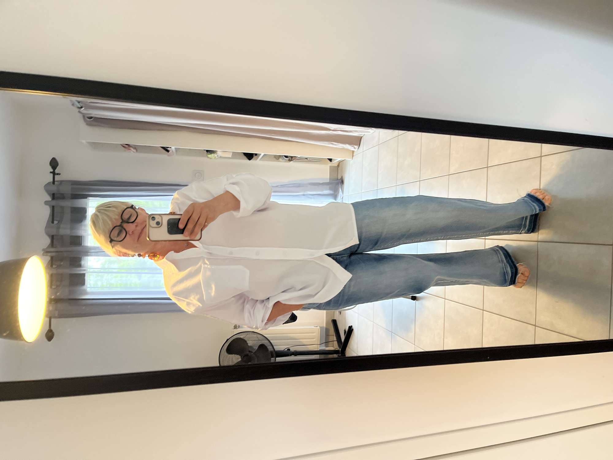 A woman with short blonde hair and eyeglasses is taking a selfie in a mirror. She is wearing a white button-down shirt and blue jeans, standing barefoot on a tiled floor. Behind her in the mirror's reflection is a room with a fan, a radiator, a window with grey curtains, and shelves with various items. The location is Chambœuf, France.