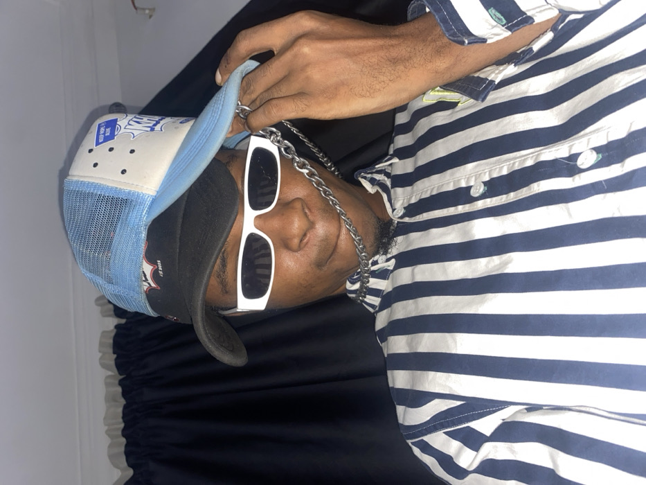A man wearing a black and light blue baseball cap, white-framed sunglasses, and a silver chain is seen. He is wearing a blue and white striped short-sleeved shirt with visible buttons. He appears to be lying on a dark, possibly black, fabric, possibly a bed. The image is lit with moderate indoor lighting.