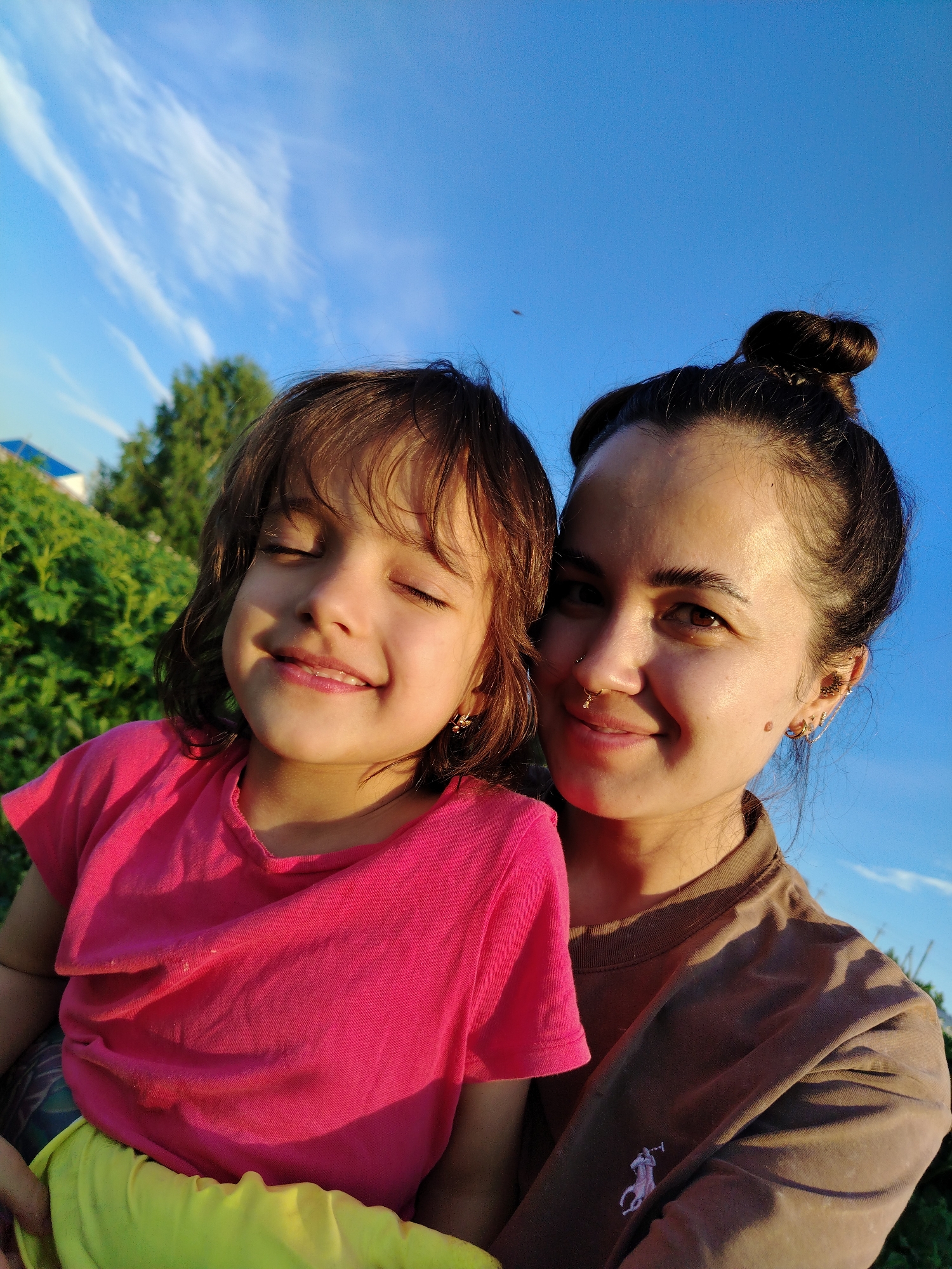A medium shot photograph captures two individuals, a young girl and an adult woman, posing for a selfie outdoors. The girl, positioned in the foreground on the left, has her eyes closed and is smiling. She wears a bright pink t-shirt and a yellow garment. To her right, the adult woman, with her hair styled in a bun, smiles at the camera. She is wearing a brown long-sleeved shirt. Both individuals are positioned against a backdrop of a clear blue sky with wispy white clouds. Green foliage and trees are visible behind them, suggesting an outdoor setting in a rural or suburban area. There are no identifiable landmarks indicating the city of Gzhan, Russia, in this image. The lighting suggests it is daytime, possibly late afternoon due to the warm glow on their faces.