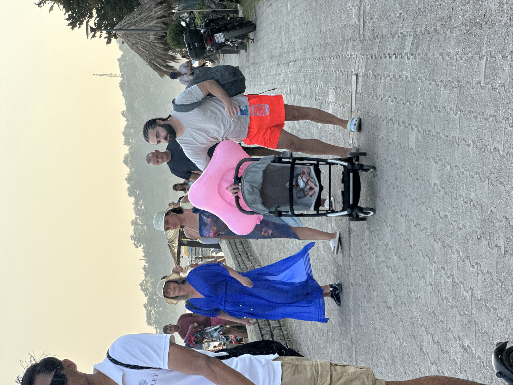 A group of people are walking along a paved walkway. In the foreground, a man with braided hair is pushing a stroller and holding a pink inflatable object. He is wearing a light grey t-shirt and red shorts. Behind him, a woman in a blue dress and a hat is walking. Further back, other individuals are visible, as well as a motorcycle parked on the right side of the walkway. In the background, there are trees and a hill with a tower structure. The walkway appears to be in a public area, possibly a promenade or park, near a body of water. The location is identified as Boreti, Montenegro.