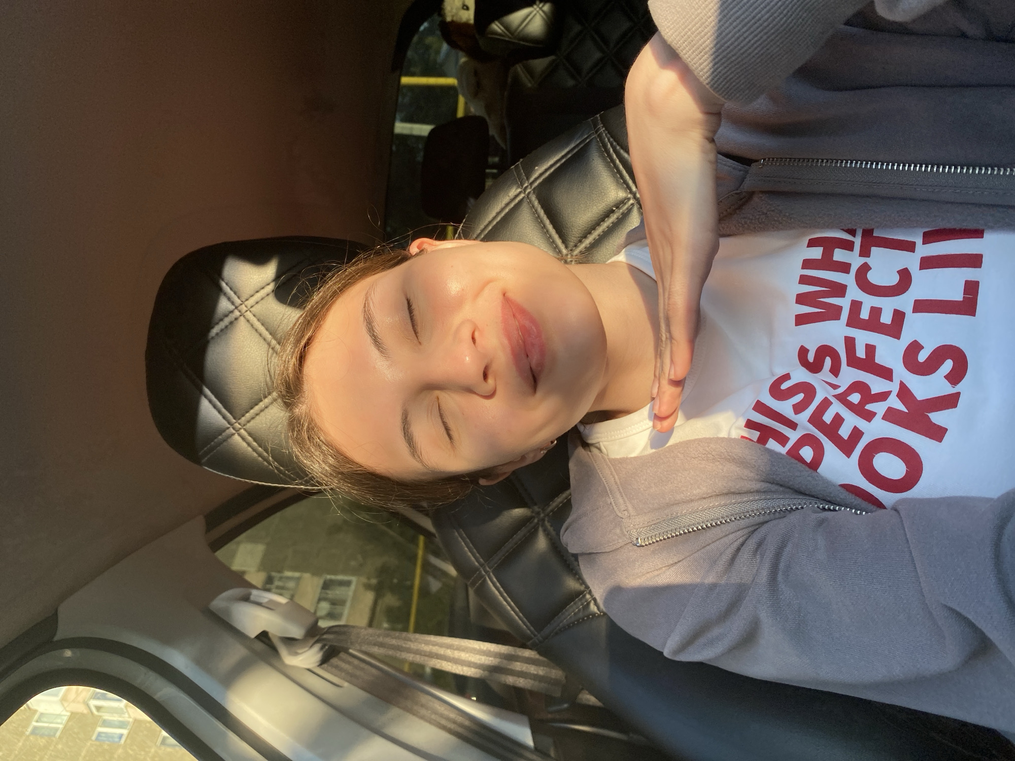 A young woman with her eyes closed is seated in the driver's seat of a car, bathed in warm sunlight. She is wearing a white t-shirt with red text that reads "THIS WHATEVER PERFECT LOOKS LIKE" and a light grey zip-up hoodie. Her right hand is gently resting on her chin. The car's interior features black leather seats with a diamond-stitched pattern. Through the car window, a building with multiple floors and windows is visible, suggesting an urban environment. The lighting indicates it is either sunrise or sunset.