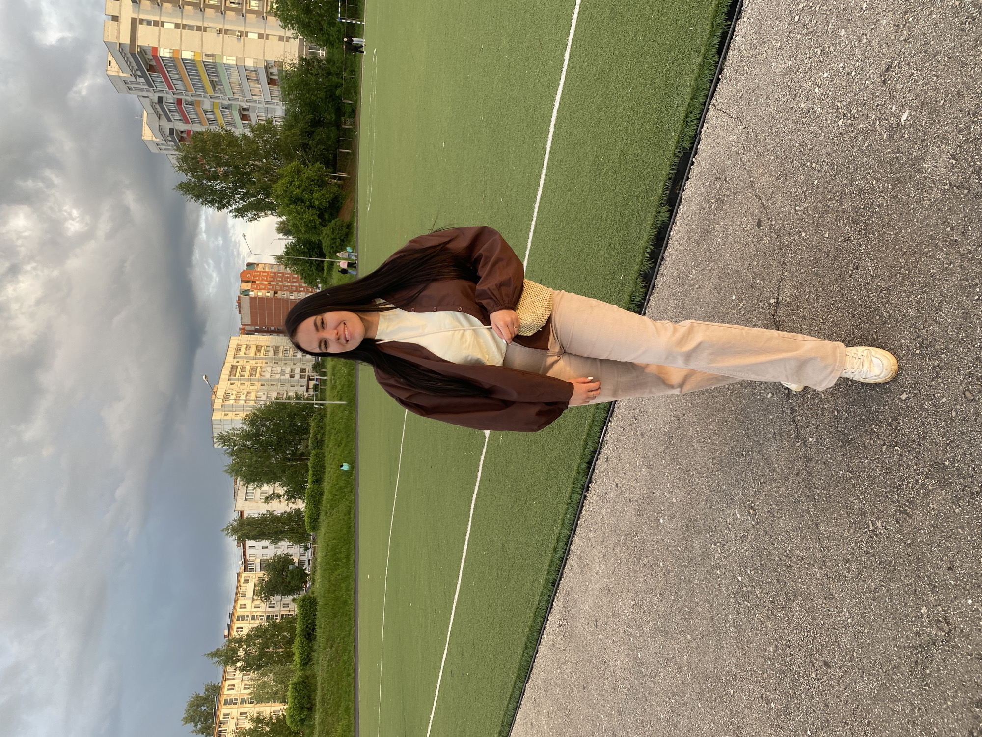 A young woman stands on a paved area beside a green sports field in what appears to be the city of Chaikovsky, Russia. She is wearing a brown jacket, light-colored pants, and white sneakers, and carrying a light-colored handbag. The background includes apartment buildings, trees, and a cloudy sky.