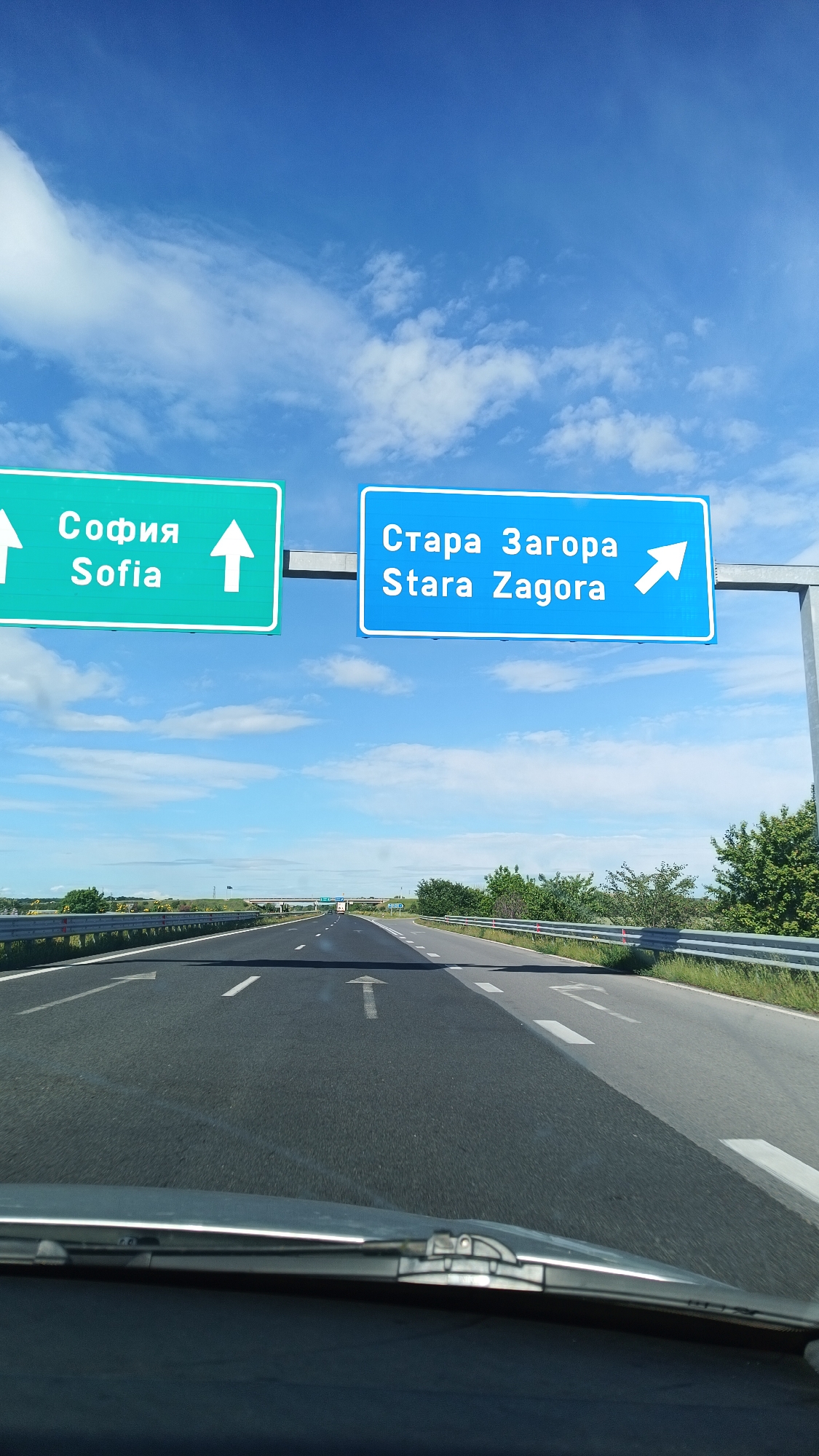 The image shows a view from a vehicle on a highway, likely in Bulgaria based on the road signs. Two prominent road signs hang overhead: one green sign points straight ahead towards "Sofia" (София), and a blue sign directs traffic to the right towards "Stara Zagora" (Стара Загора). The sky is blue with scattered clouds. The highway has multiple lanes with white directional arrows painted on the asphalt. The immediate foreground shows the vehicle's dashboard and windshield. The highway stretches into the distance, bordered by green vegetation on the sides.