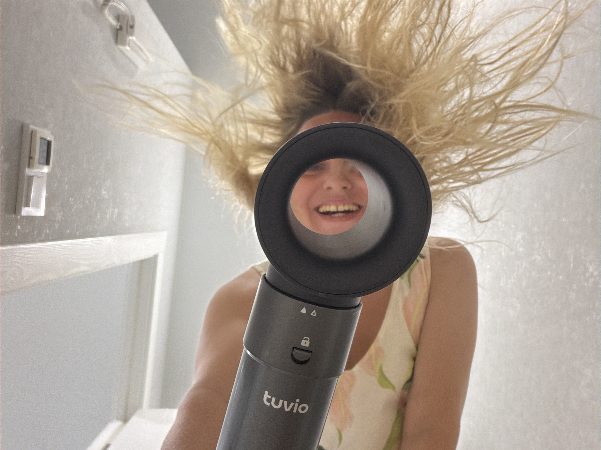 A low-angle, medium shot shows a woman holding a black hairdryer, with the nozzle pointed towards the camera. Her blonde hair is blown upward by the airflow, creating a wild, voluminous effect that fills the top of the frame. She is smiling broadly, with her mouth open, and appears to be enjoying the experience. The woman is wearing a light-colored sleeveless dress with a floral pattern. The background is a softly lit, pale textured wall, with some faint light fixtures visible on the left side of the image. The hairdryer has "tuvio" written on its body, along with a lock symbol and two triangle symbols. The overall impression is one of playful exuberance and carefree fun. The time of day and weather are not discernible from the image. The location is Votkinsk, Russia.