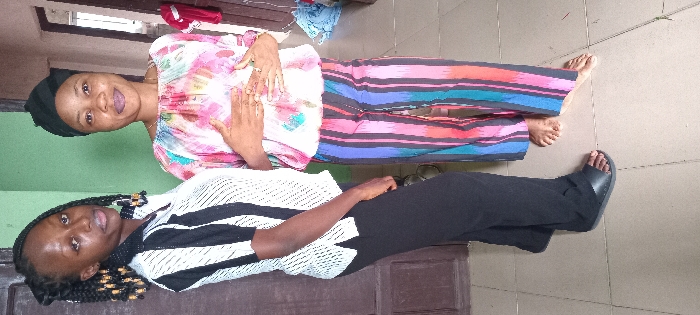 Two women are posing for a picture. The woman on top has on a pink and white top and colorful pants. She is wearing a black head covering. The woman on the bottom has on a white and black striped top and black pants. She is wearing black sandals. The women are inside a house with linoleum floors.