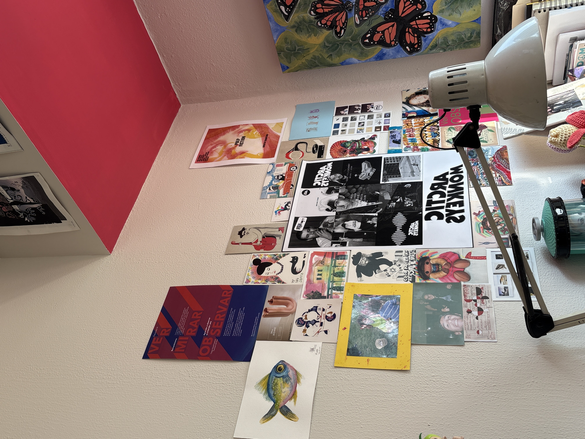 The image shows a section of a wall covered with various posters and artwork. The wall is painted white, except for a portion at the upper left which is a bright red. The posters include images of musical artists such as the Arctic Monkeys and Pulp Fiction, as well as various graphic designs and photographs. A desk lamp is positioned on the right side of the frame, its head pointing towards the wall. The scene appears to be in a room in Jesús del Monte, Mexico, based on the text "Jaffa" and "DITORA" and the local aesthetic. There are no people visible.