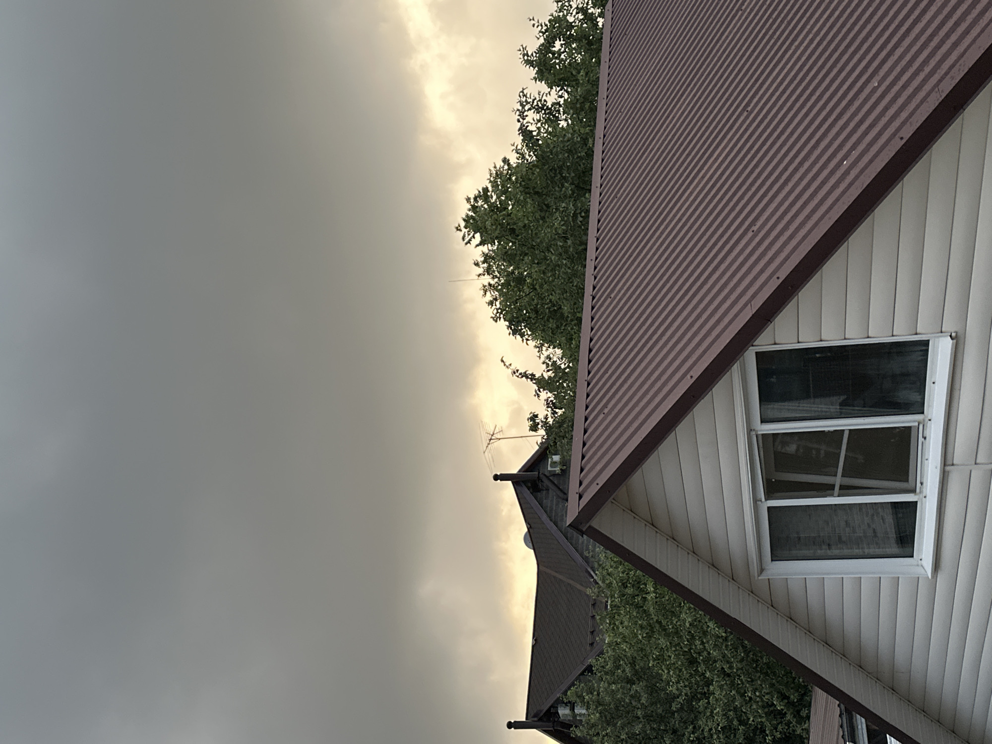 The image captures a residential scene with multiple houses and trees under a cloudy sky. In the foreground, a house with a beige, horizontal siding and a dark brown, corrugated metal roof is visible. The house has a white-framed window with multiple panes, some of which are open. Behind this house, another house with a dark brown, shingled roof and dark brick exterior is partially obscured by lush green trees. This house has two visible white air conditioning units and a satellite dish on its roof. Further in the background, the outline of another house with a dark roof is visible. A television antenna stands tall against the overcast sky. The overall atmosphere suggests a potentially overcast or approaching storm weather. The scene is typical of a suburban or rural residential area, possibly in Nizhny Novgorod, Russia, given its geographic characteristics and architectural styles commonly found in such regions. There are no people or animals present in the image.