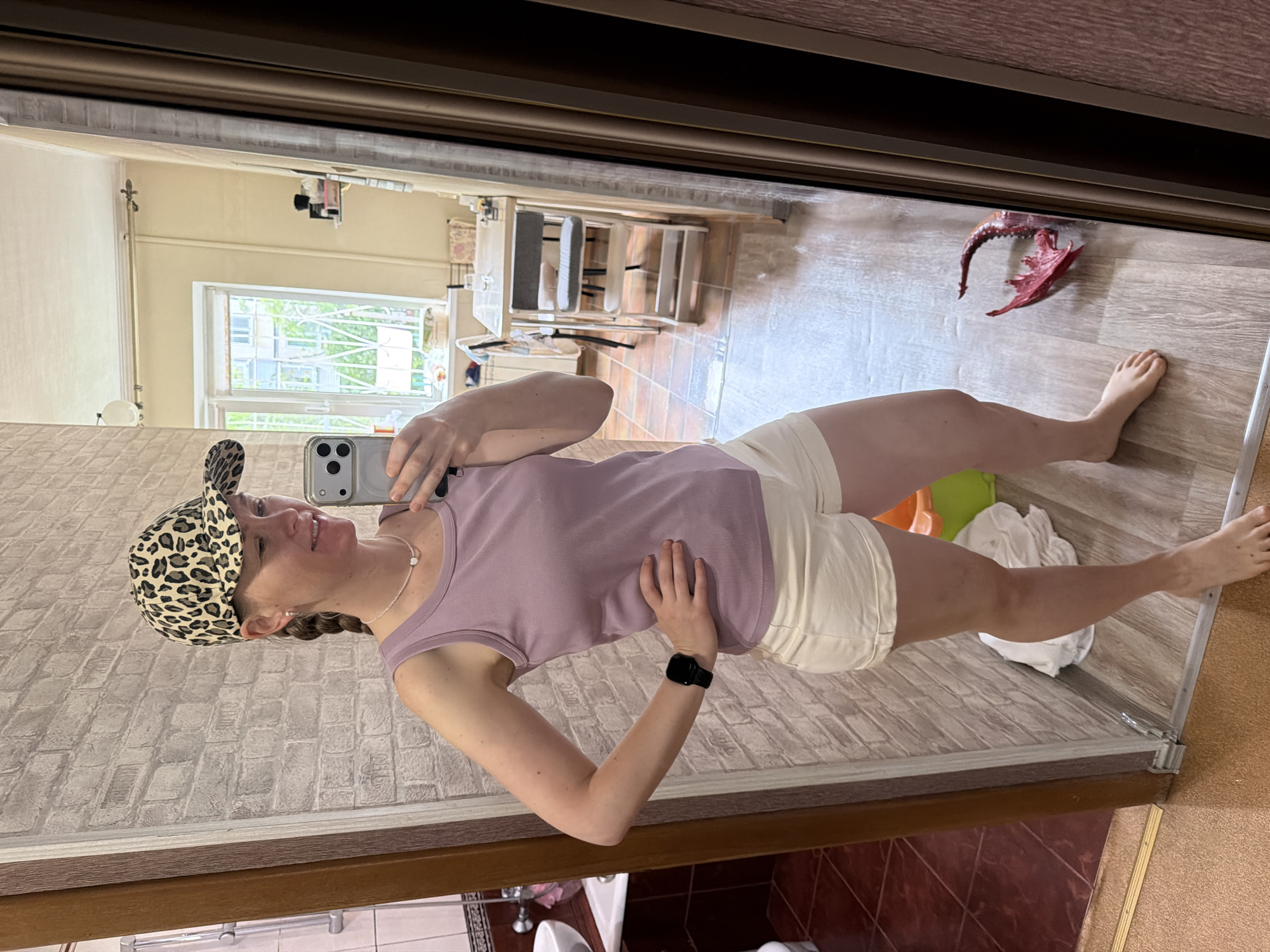 A young woman stands in a room, taking a selfie in a mirror. She is wearing a light purple tank top, cream-colored shorts, and a leopard-print baseball cap. She has a braided pigtail and is wearing a delicate necklace and a black smartwatch. Her left hand is on her hip, and she is holding a smartphone in her right hand.
The room appears to be an apartment or house. The background shows a kitchen area with a table and chairs, a window with a view of trees and buildings outside, and a tiled bathroom area visible to the left. There are also some household items on the floor, including a white cloth and a bright orange bucket. A decorative dragon tail sculpture is visible on the floor to the right.
The image does not contain any specific visual indicators to place it in Khabarovsk, Russia.