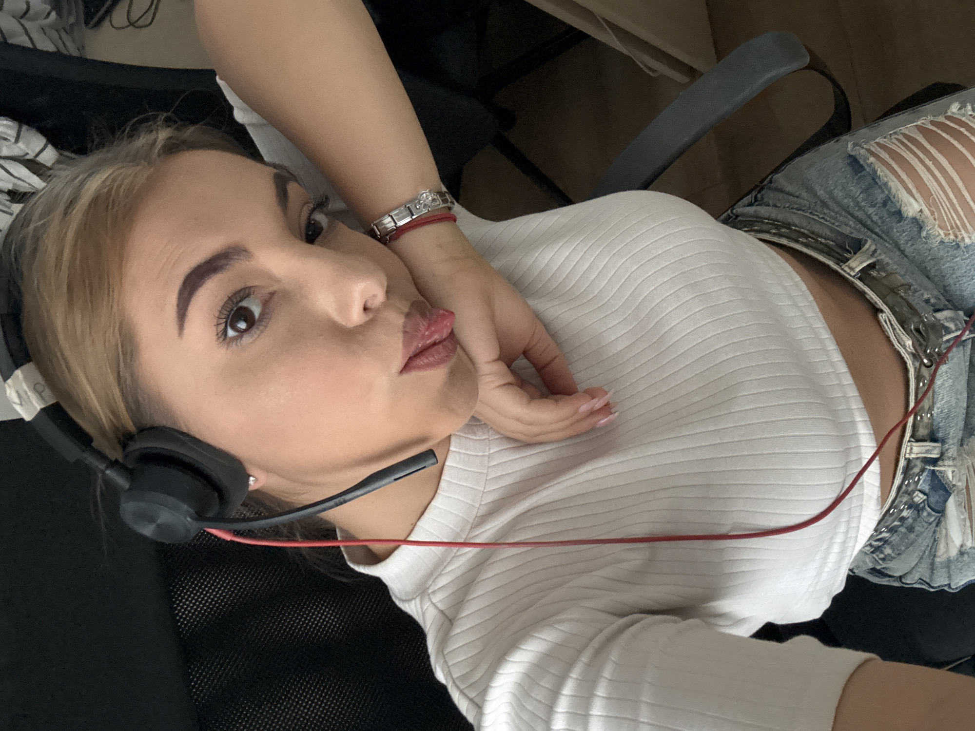 The image shows a young woman wearing headphones, a white ribbed crop top, and ripped denim shorts. She is sitting in a black chair in what appears to be an indoor setting. Her left arm is resting on her cheek, and she has her tongue sticking out slightly. A red cord from the headphones is visible, extending down her torso. She is wearing a silver bracelet on her left wrist with a red string attached. The background is dark and out of focus, with some hints of furniture and what might be a patterned fabric on the right side. There is no indication of the specific location within the city of Sofia, Bulgaria.