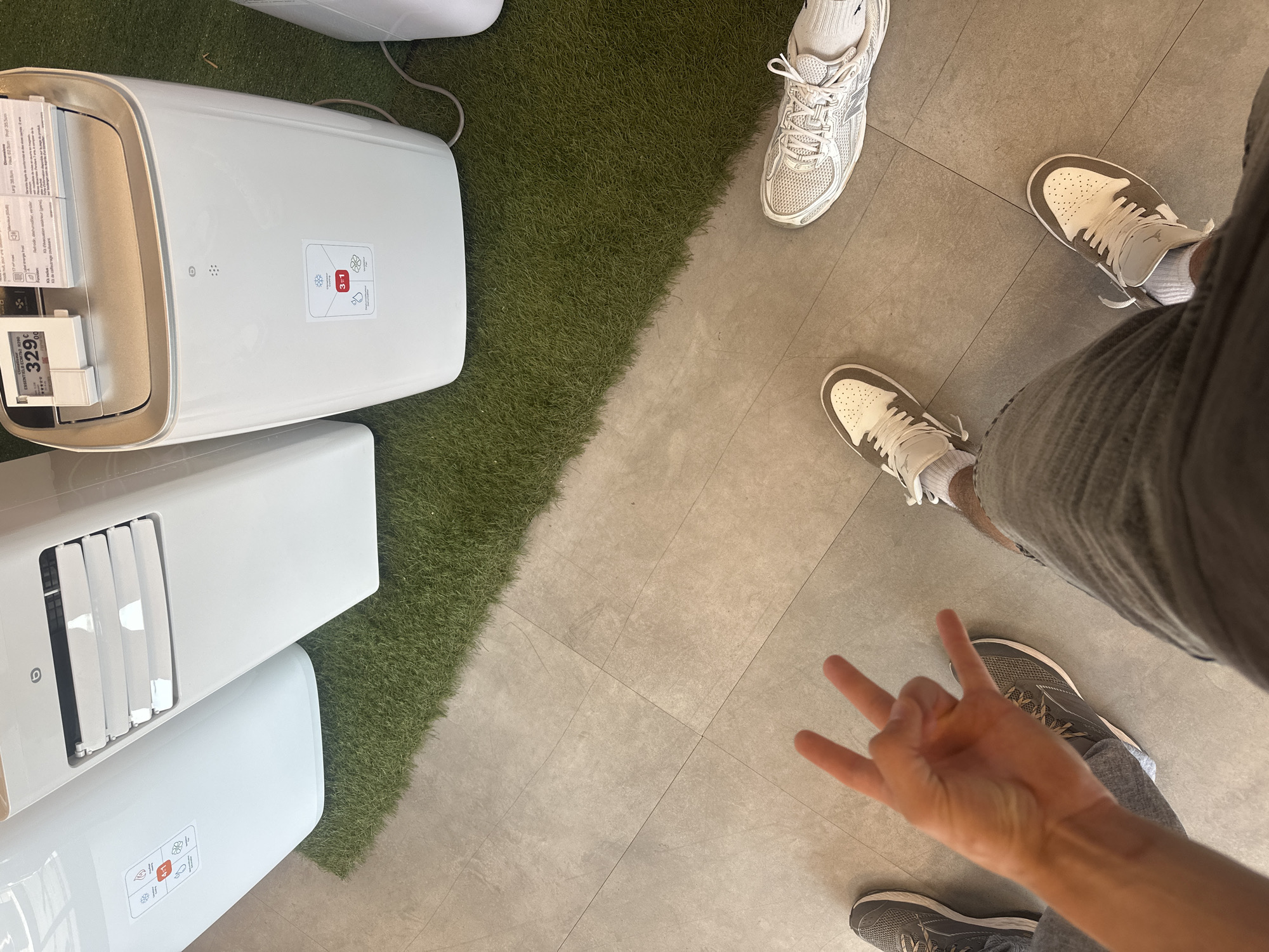 This is a top-down, wide-angle photograph taken indoors. The scene features two individuals, indicated by their visible footwear. One person is wearing white sneakers with grey accents and white socks. The other person is wearing grey sneakers with white laces and soles, and grey pants.
The ground is a mix of grey tiled flooring and a section of artificial green turf, creating a distinct visual divide. On the turf, there are several white appliances, possibly portable air conditioners or dehumidifiers, stacked vertically and horizontally. One appliance is prominently displayed with a price tag reading "329.00" and "ESSENTIALS HOME". Another appliance has a label with icons indicating functions like cooling, heating, and dehumidifying, along with a "3-in-1" designation.
One of the individuals has their hand in the foreground, with fingers extended in a gesture that could be interpreted as "peace" or a number. The lighting appears to be artificial, suggesting an indoor setting like a retail store or showroom. There are no obvious indications of the time of day or weather outside. The overall impression is a casual snapshot, possibly taken while shopping or browsing.