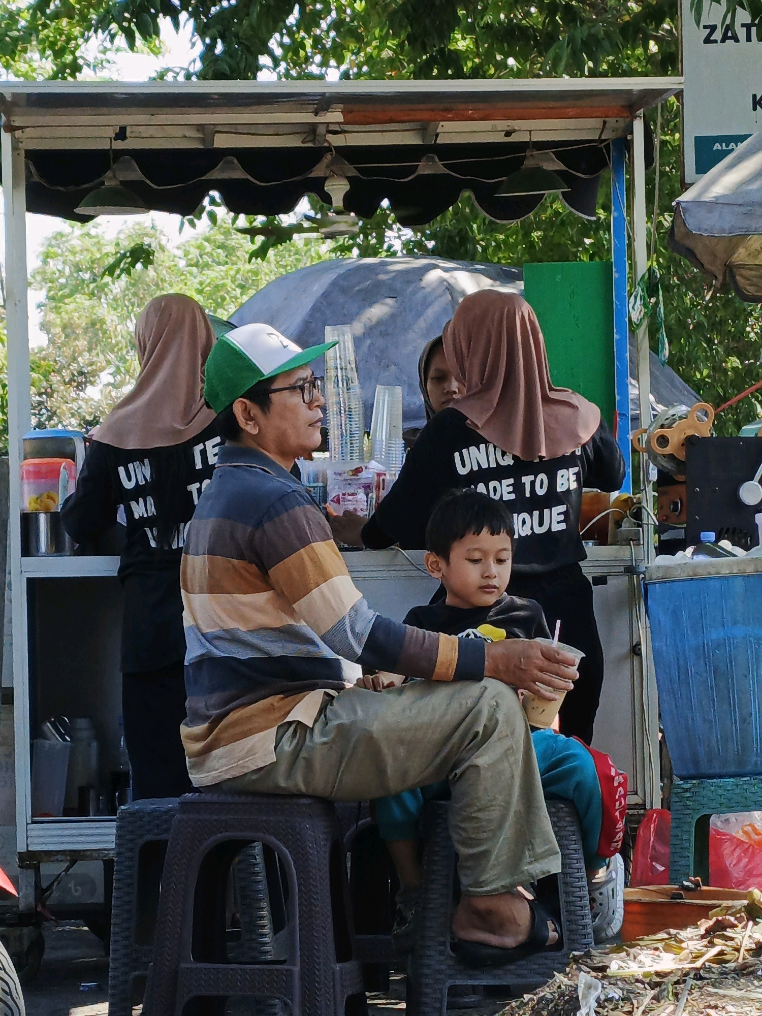 In Sumokali, Indonesia, an outdoor food stall features a man wearing glasses, a striped long-sleeved shirt, and a green and white baseball cap, seated on a dark plastic stool. He holds a plastic cup with a straw. Beside him, a young boy in a black shirt sits on a similar stool. Behind them, two women in hijabs are working at the stall. One woman wears a black shirt with white text that reads "UNIQUE MADE TO BE UNIQUE", and the other wears a black shirt with white text that is partially visible. The stall is equipped with a counter, cups, and what appears to be a juice press. Trees and foliage are visible in the background, indicating an outdoor setting.