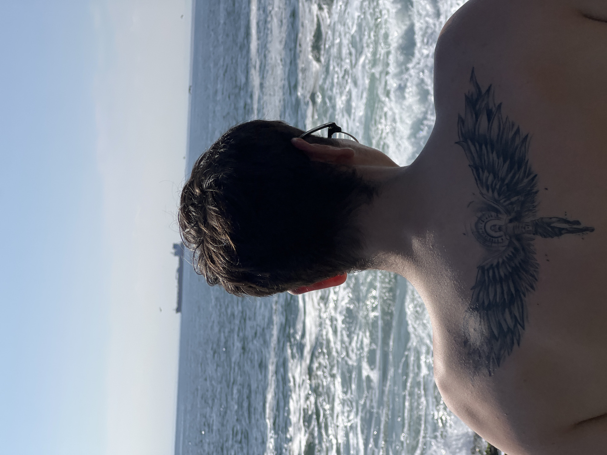 A person with dark curly hair and a tattoo of wings on their back stands facing the ocean. They are wearing glasses. The water is choppy with white foam. In the distance, a ship is visible on the horizon. The location is most likely on the coast of Arica, Chile, given the context.