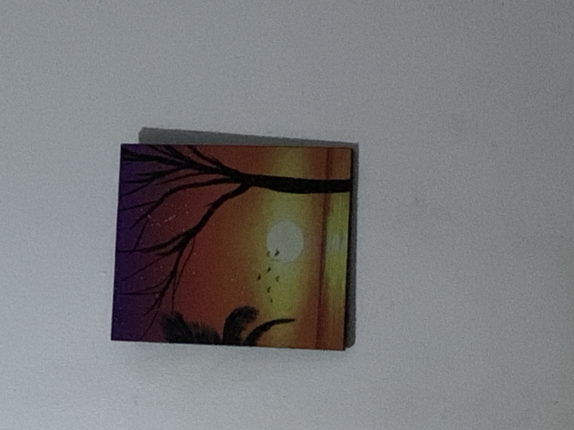 A small, square painting of a sunset scene is affixed to a plain, light-colored wall. The painting depicts a dark silhouette of a tree with bare branches against a gradient sky of deep purple at the top, transitioning to orange and yellow towards the horizon. A bright, pale circle, representing the sun, is visible near the horizon. A few small, dark shapes in the sky suggest birds in flight. To the left of the tree, a dark silhouette of a palm frond is partially visible. The foreground of the painting shows a body of water with a reflective surface mirroring the colors of the sky. The wall is textured and appears to be a typical interior surface. The location in Barakin Lerawi, Nigeria, is not discernable from the image itself.
