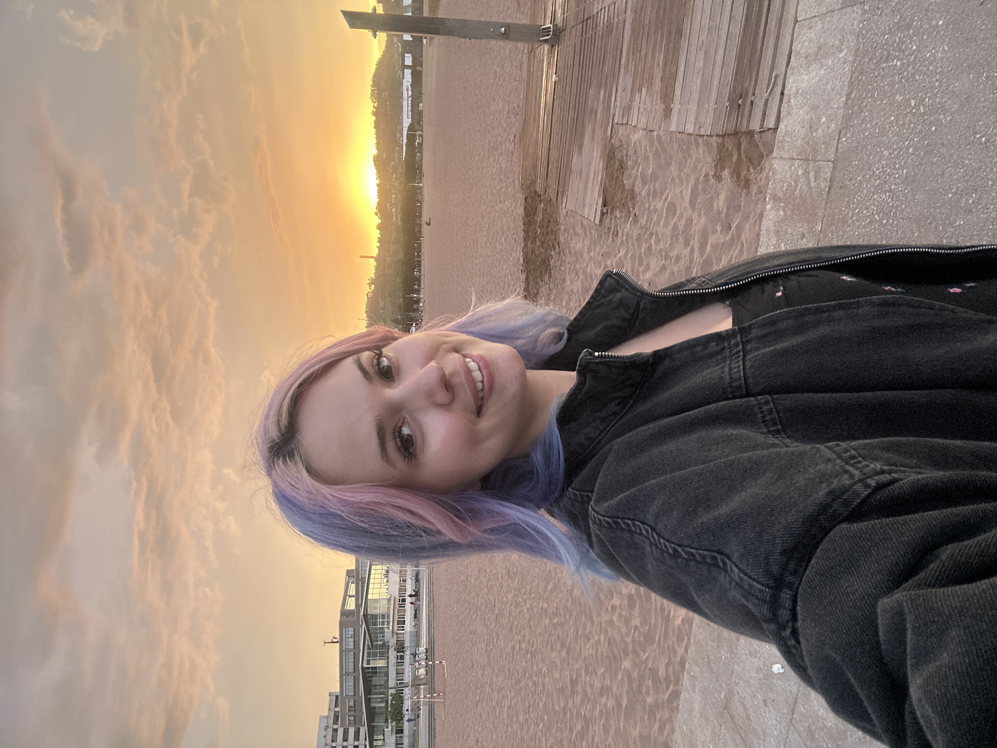 A young woman with pink and blue dyed hair takes a selfie on a beach in Gijón, Spain. She is wearing a dark denim jacket over a black top. The background shows a sandy beach with a wooden boardwalk to the right. In the distance, there are buildings and hills, and the sun is setting, casting a warm orange glow across the sky. The overall scene is serene and depicts a casual moment at the beach.