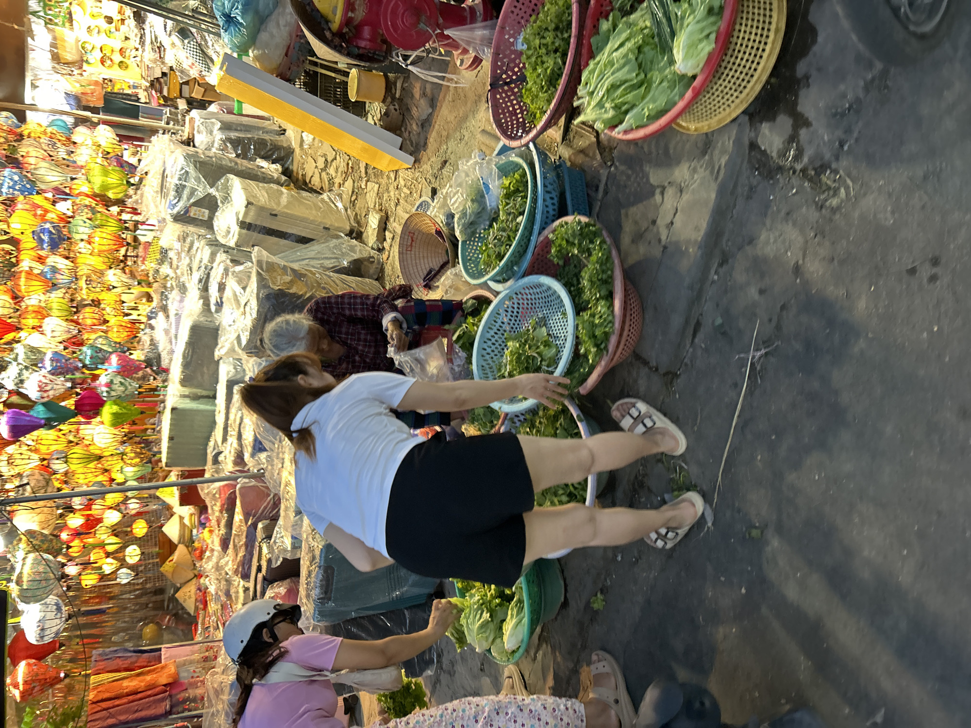 In Hội An Ward, Vietnam, a street vendor scene unfolds at dusk, illuminated by numerous colorful lanterns hanging overhead. A young woman in a pink shirt and patterned pants, wearing a white helmet, is seen from the side, bending down to examine produce. To her right, a woman in a white t-shirt and black shorts is also bent over, sorting through green vegetables in a basket. Behind them, an elderly woman in a plaid shirt and dark pants sits, seemingly engaged in packing or arranging items. The ground is paved with asphalt, showing signs of dampness and reflections from the lanterns. Various baskets filled with fresh green vegetables, including lettuce and leafy greens, are displayed on the ground. In the background, stacks of items covered in plastic sheeting and what appear to be suitcases are visible, suggesting a market or shop setting. A bright yellow and white sign stands vertically in the mid-ground, and a red fire hydrant with a yellow bucket nearby is present on the right side of the image. The overall atmosphere is that of a bustling evening market.