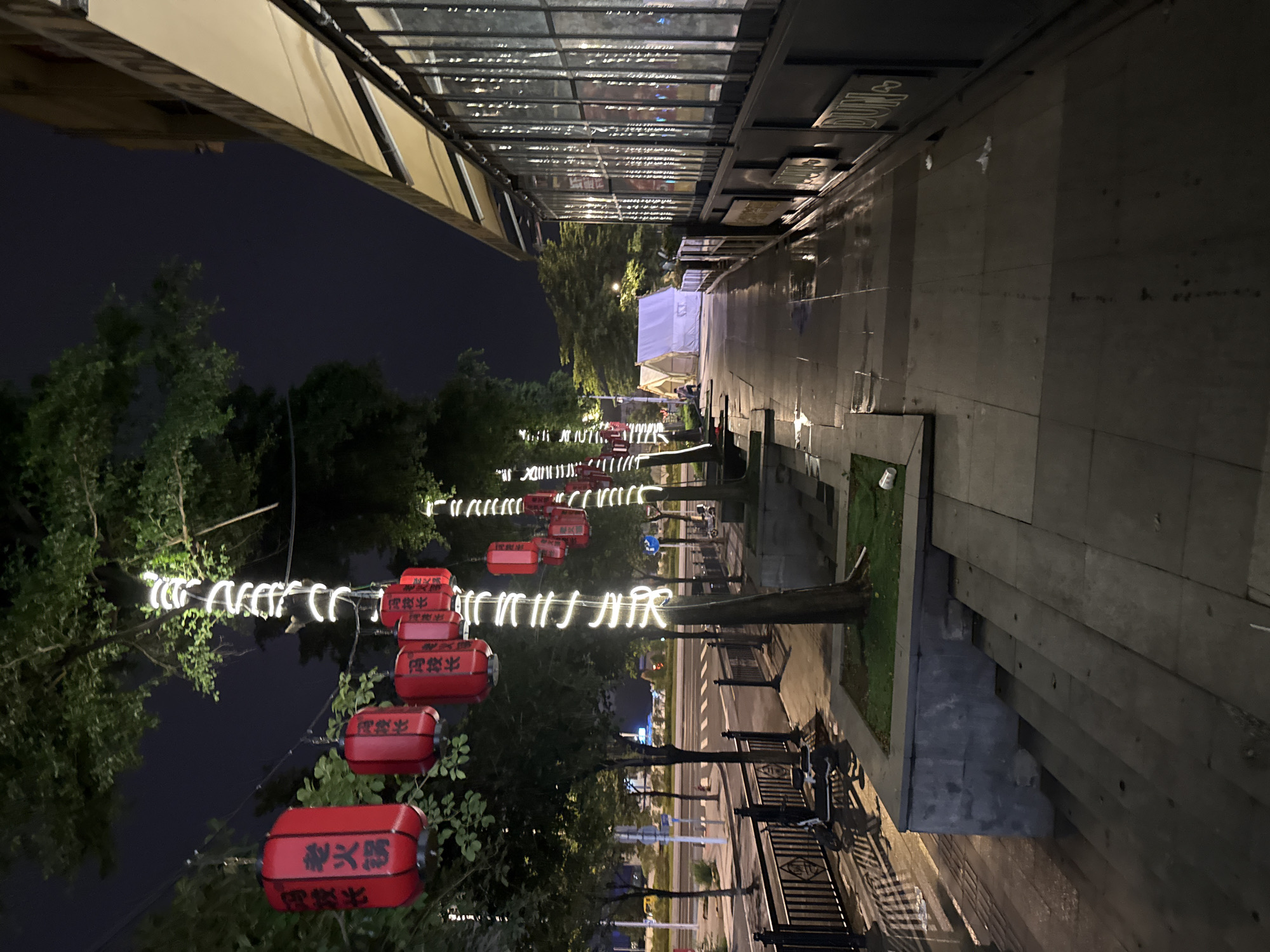 The image captures a nighttime street scene in Haizhu District, China. The foreground features a concrete walkway with stairs leading down to a street. The walkway is lined with trees decorated with white string lights. Red lanterns with Chinese characters are hung from wires above the walkway. To the right of the walkway is a building with large windows, partially covered by awnings, and signs with English text, including "DUN". In the background, a street with traffic lights and a few vehicles is visible. Further back, there is a white tent structure. The ground on the walkway appears wet, suggesting recent rain. The overall impression is of a well-lit, commercial area at night.