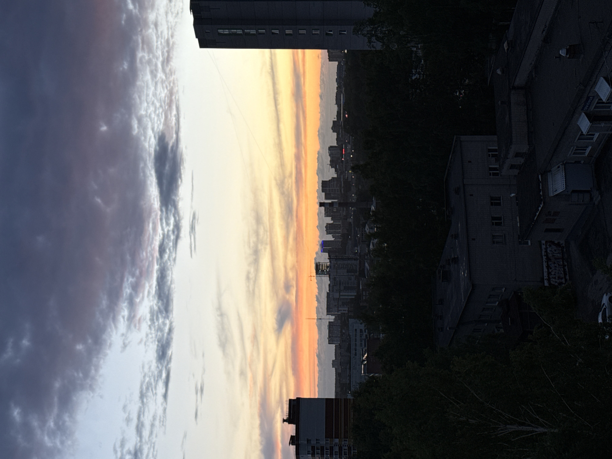 The image shows a sunset over the city of Izhevsk, Russia. The sky is filled with clouds, with shades of orange, pink, and gray. Below the sky is a cityscape with buildings of varying heights. In the foreground are trees and parts of buildings. Some buildings show windows. The scene appears to be taken from an elevated position.