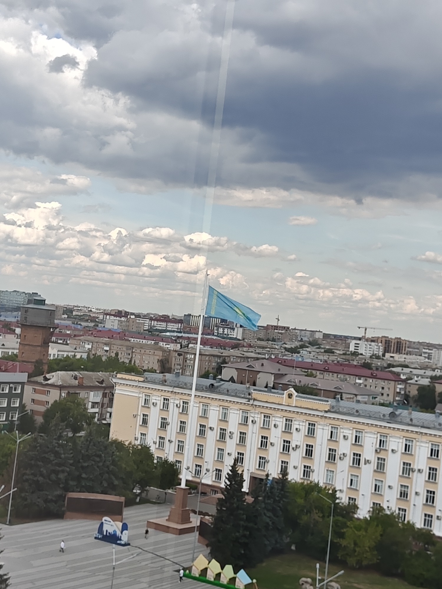 The image shows an aerial view of a city square in Petropavl, Kazakhstan, under a cloudy sky. A prominent feature is a large, light-colored building with many windows, suggesting it is a public or administrative building. In front of this building, a flag of Kazakhstan is flying on a tall flagpole. To the left of the building, a tall, brick structure, possibly a water tower, is visible. The square itself is paved, with a monument or memorial in the center and some trees and greenery bordering the area. A few people are present in the square, walking or standing, and some small, colorful structures are also scattered around. The surrounding cityscape consists of numerous residential and commercial buildings of varying heights and architectural styles, indicative of an urban environment. The overall impression is a civic space within a city.