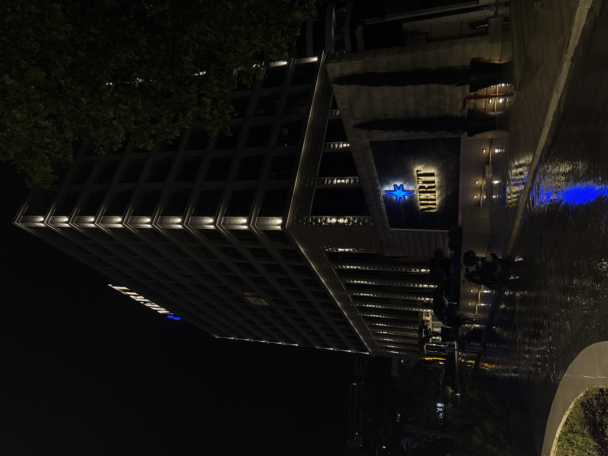 This is a night shot of the Merit Casino in Budva, Montenegro. The building is tall and angular, with illuminated edges. The Merit logo, a blue star, is displayed prominently both on the side and at the entrance. A water feature runs along the entrance, reflecting blue light. Several scooters are parked in front of the entrance. There is a tree on the left side of the frame.