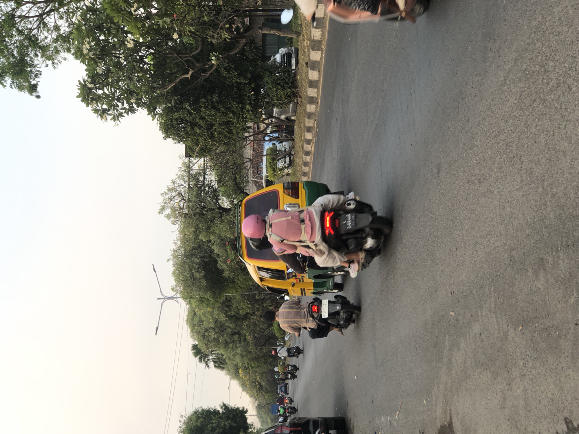 This is a daytime, eye-level shot of a street in Ngaban, Indonesia, with moderate traffic. In the foreground, a yellow and green van is driving on the right side of the road. Behind the van, two people are riding on a black scooter. The person in the back is wearing a pink helmet and a pink shirt. The person in front is wearing a patterned shirt. To the left of the van, there are several other scooters and a black car driving in the same direction. Further down the road, there are more scooters and trees lining the street. The sky is overcast, and there is a street lamp visible in the distance. The road surface is asphalt, and there is a concrete curb on the right side of the road.