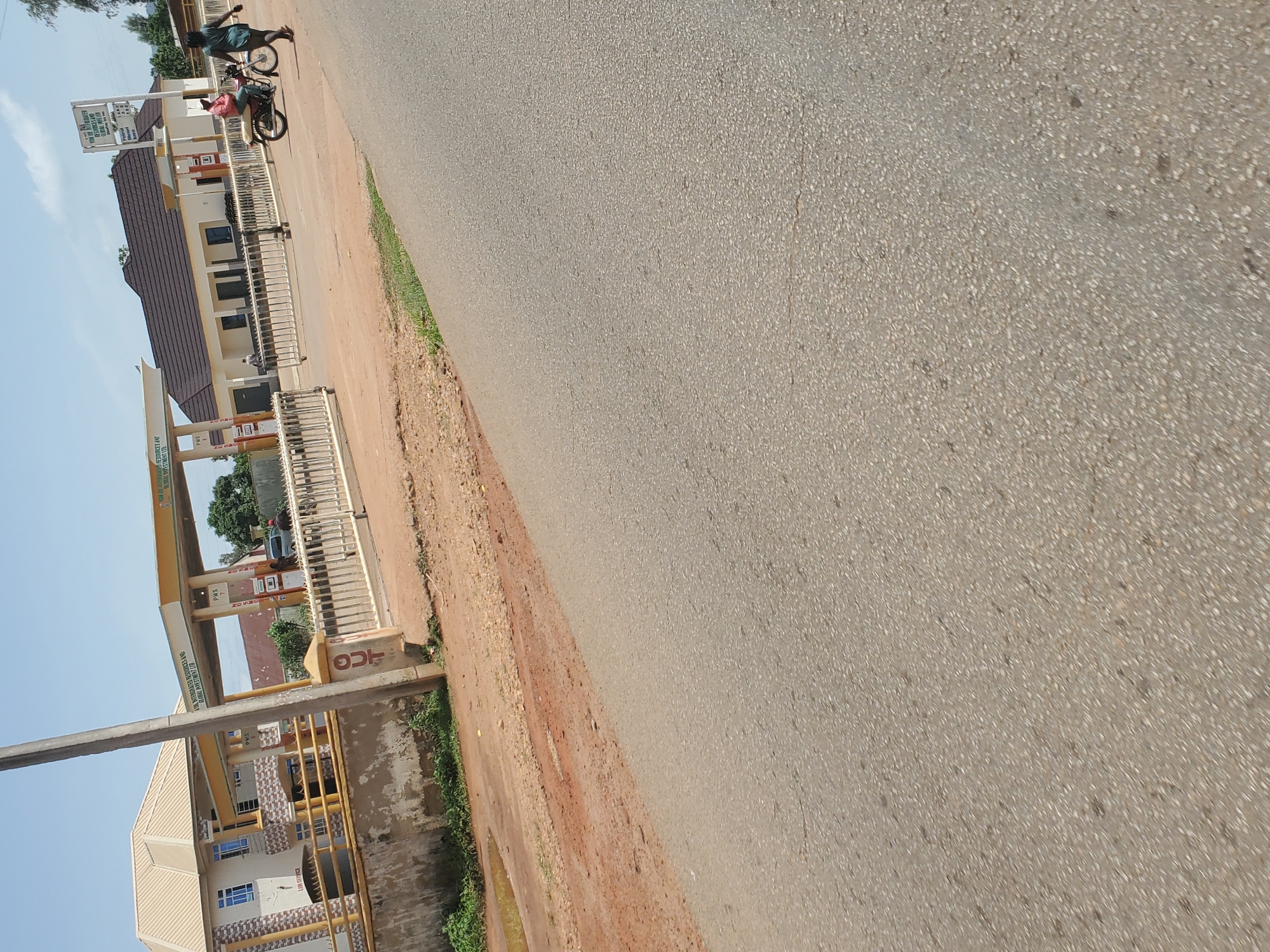 The image, taken in Vom, Nigeria, depicts a daytime roadside scene featuring a fuel station adjacent to a paved road under a clear, bright sky. The fuel station, prominently on the left, is identified by a yellow and brown canopy structure. A large vertical white sign displays the name "D. O. G. S. ENTERPRISES LIMITED" and advertises "Quality ASSURED QUANTITY," along with "OUR CORE VALUES" listed as Safety, Integrity, Customer Focus, Excellence, and Teamwork. Another partial sign on a canopy support structure reads "D. O. G. S. ENERGY RESOURCES AND INVESTMENT LIMITED."
Beneath the canopy, several fuel pumps are visible, indicating "PMS" (Premium Motor Spirit) and "DIESEL," with "N0.00" displayed for prices. A building with a brown roof and light-colored walls stands behind the pumps, separated by a white metal fence or railing. Two individuals are near the fuel station's entrance area. One person, wearing a dark shirt and dark trousers, is standing next to a motorcycle or scooter. The second individual, dressed in a red top and dark trousers, is seated on the same motorcycle. Their activity suggests a stop at the fuel station, possibly for refueling or a break.
The road surface in the foreground and stretching into the distance is grey asphalt. Alongside the road is a wide, unpaved shoulder of reddish-brown dirt and gravel, which transitions into patches of green grass and vegetation. A grey utility pole stands upright in the lower-left foreground. A concrete pillar or block with the letters "JCG" is also visible near the roadside. The bright lighting indicates the time of day is likely late morning or afternoon.