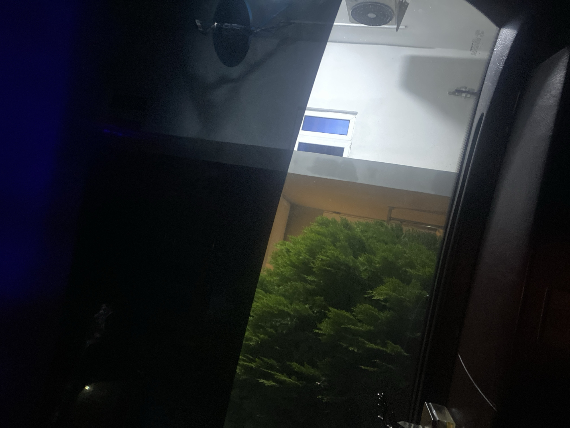 The image depicts a view from inside a vehicle looking out at night. In the foreground, the dark interior of the car is visible, with a portion of the dashboard and windshield frame. Through the windshield, a dense, green shrub is prominent on the left side. To the right of the shrub, a building with a white facade and a window with blue panes is visible. An air conditioning unit is mounted on the exterior wall of the building. The scene is illuminated by artificial light, suggesting it is nighttime. The specific location in Barikin Jirga Summa, Nigeria, is not discernible from the visual information presented in the image.