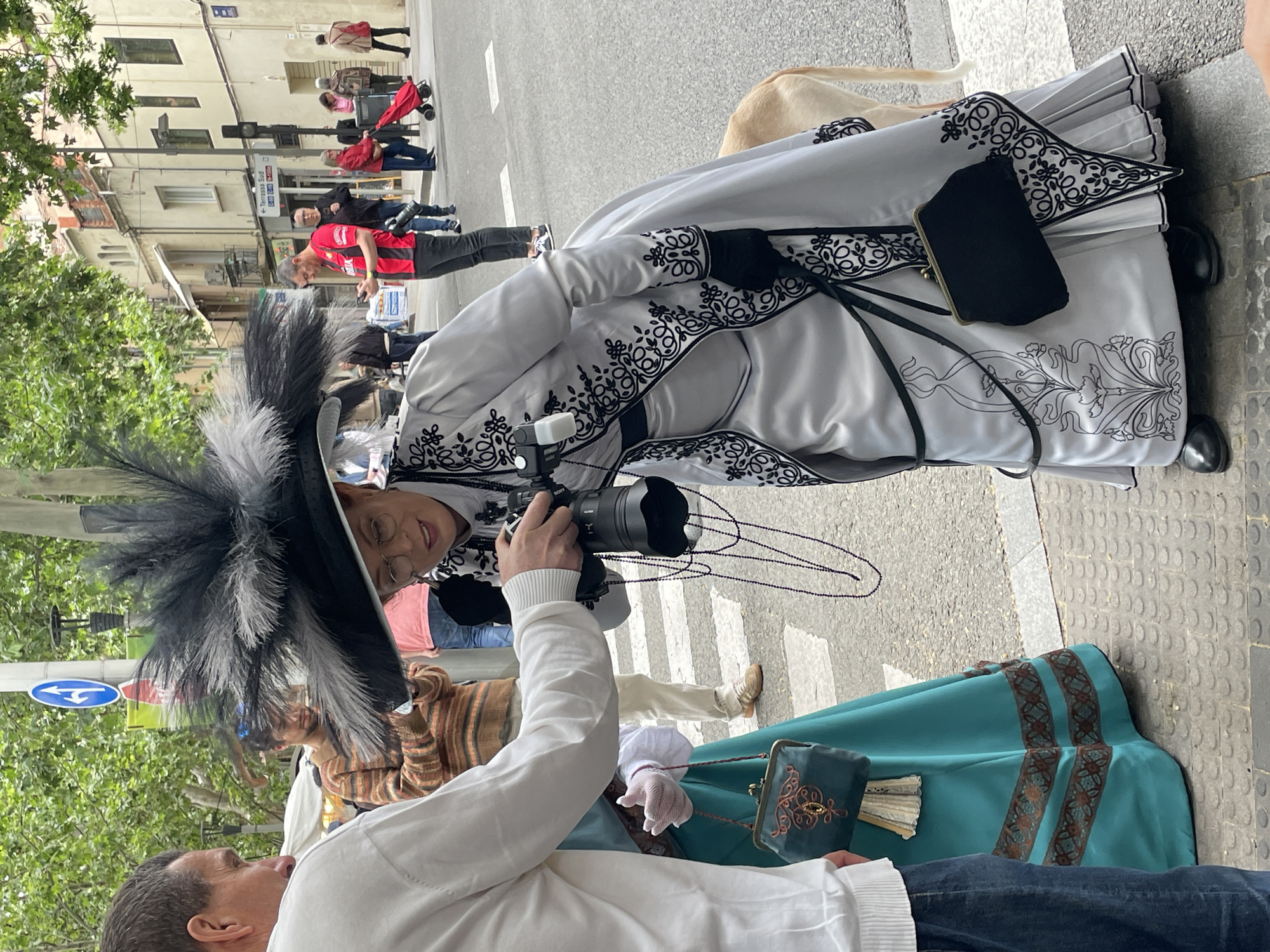 The image depicts a street scene in Terrassa, Spain, with pedestrians walking in the background. In the foreground, two individuals are interacting. One person, likely a photographer, is holding a Sony camera and is poised to take a picture. The other person is wearing a striking white and black period costume with elaborate embroidery and a large feathered hat, a small dark purse, and a long pleated skirt. A fan is visible near them. A blue and green dress of a similar period is slightly visible. The interaction takes place on a pedestrian crossing, and the surroundings include buildings and greenery, typical of an urban setting in Terrassa.