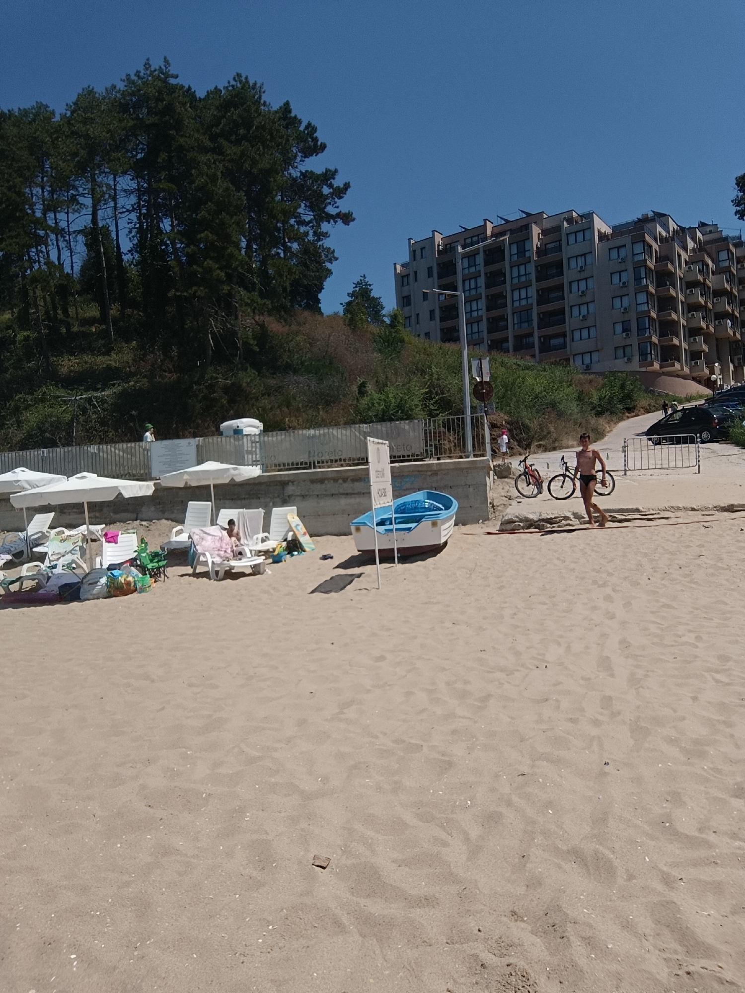 This is a daytime, eye-level shot of a beach in Obzor, Bulgaria, with a clear blue sky. In the foreground, a sandy beach is visible with several white lounge chairs, some with towels draped over them, and beach umbrellas. A small blue boat is resting on the sand. Further back, a man in dark swim trunks runs across the sand. Two bicycles are parked nearby. Behind the beach, there is a retaining wall with a metal fence, and beyond that, a multi-story modern apartment building is situated on a hill covered with trees. To the left, more pine trees cover the hillside. The overall scene depicts a typical beach day with recreational activities and resort infrastructure.