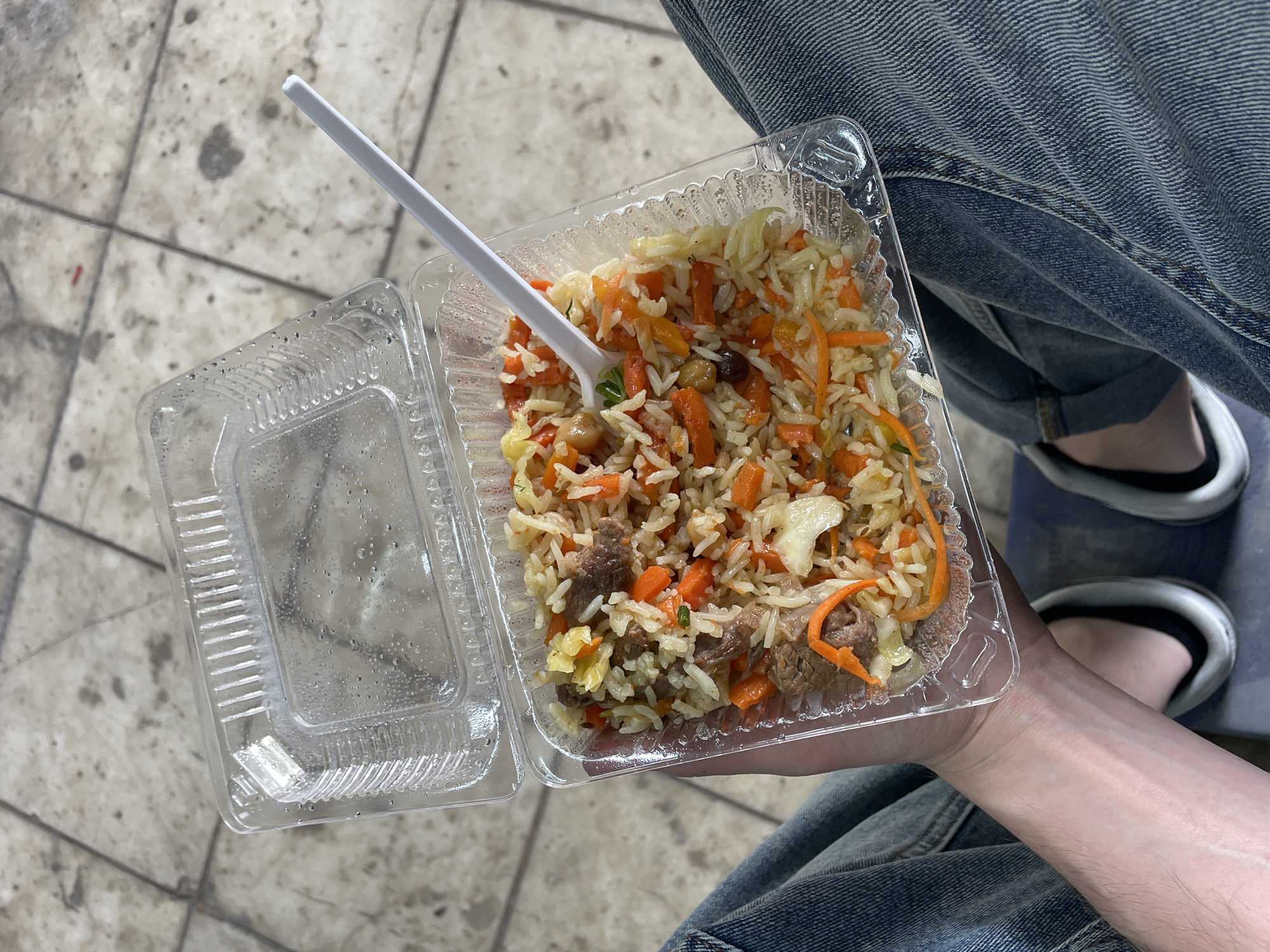 A high-angle shot depicts a person holding a partially opened, square, clear plastic container filled with cooked rice, carrots, and what appears to be meat. A white plastic fork is partially inserted into the food. The person is wearing blue jeans, and the bottom of a shoe is visible. The location appears to be an outdoor area with light-colored pavement. The scene takes place in Минино, Russia.